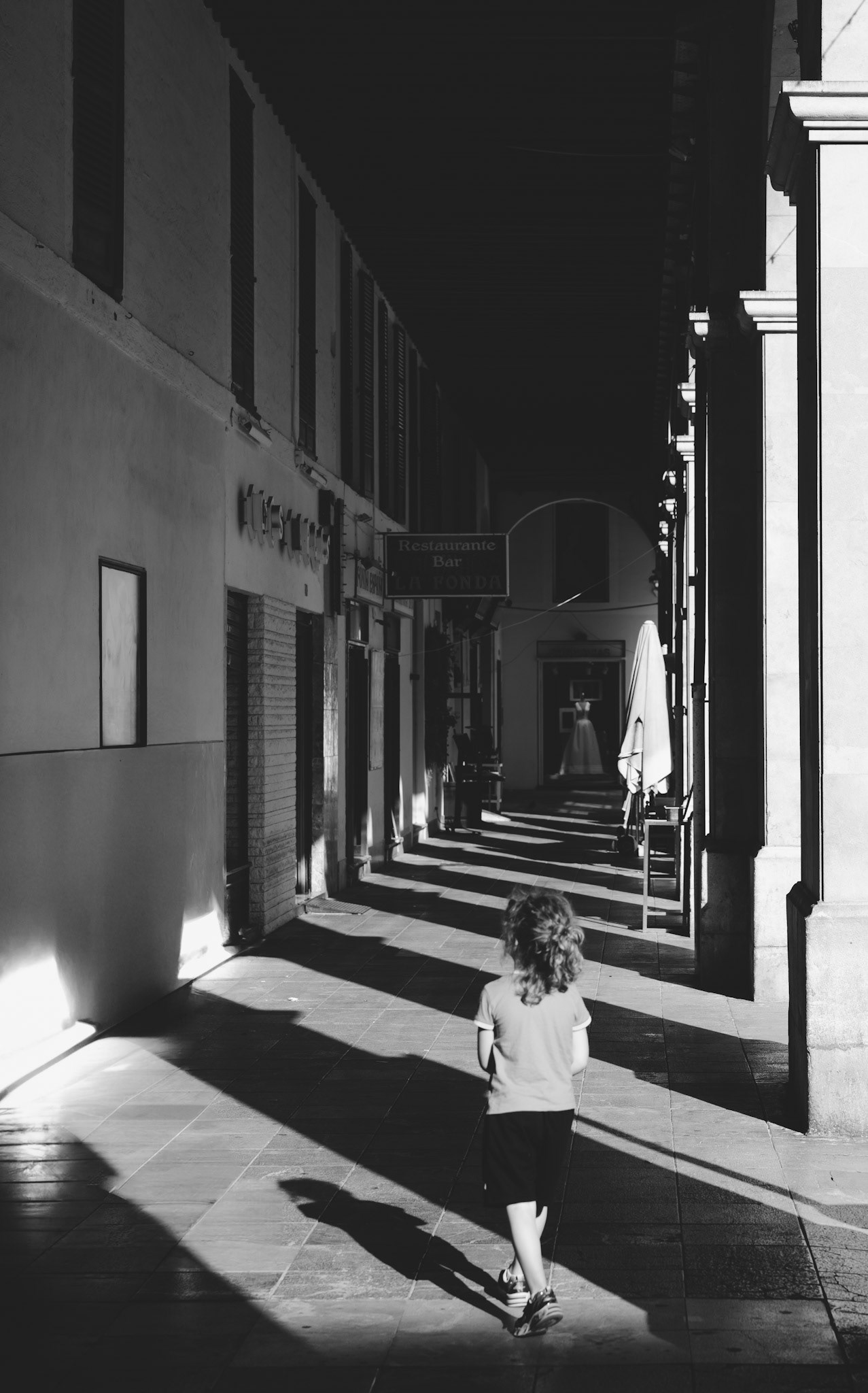 captured at plaça major in palma de mallorca, this evocative photograph captures a young child walking through the arches, casting long shadows that blend into the darkness. the play of light and shadow creates a dramatic contrast, emphasizing the feeling of being lost amidst the historic arcade. the monochrome palette adds a timeless quality to the image, highlighting the textures and details of the architecture.