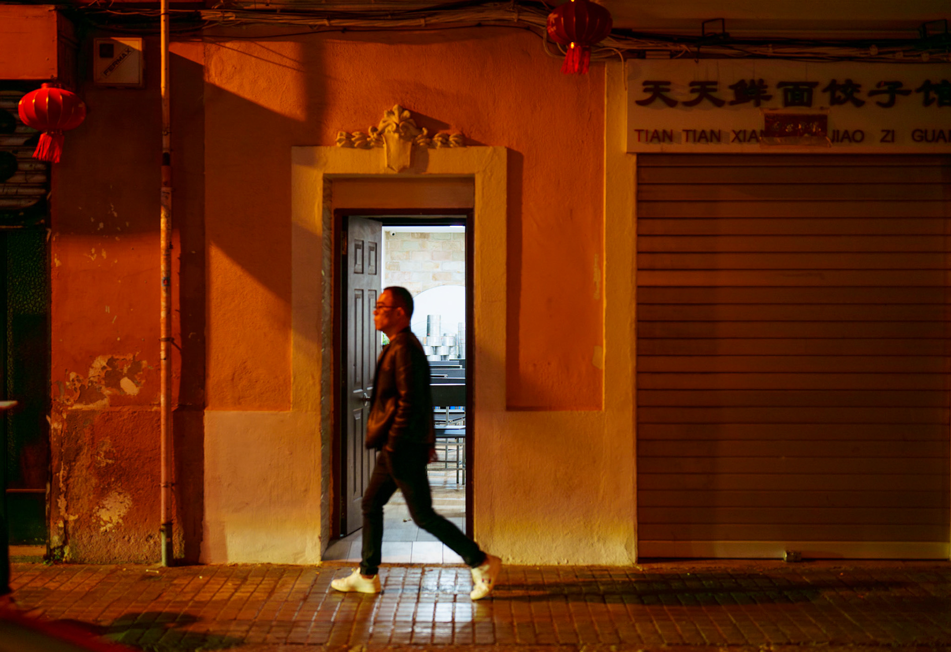 in the warm embrace of valencia's extramurs, a historical palette mingles with the zest of asian vitality along c/ de pelai. it's here where the stories, scents, and flavors of distant lands converge through doorways that serve as portals to other worlds. as evening descends, a lone figure strides across one such threshold, momentarily framed by the soft glow of ambient light that spills out onto the cobblestones. like a scene from a nocturne, every element here whispers of transitions: the old walls bearing the passage of time, the red lantern a beacon of culture, and the shuttered storefronts a nod to the restful city. this is a snapshot of life's continuous flow, the fleeting interplay of past and present, captured in a single stride.