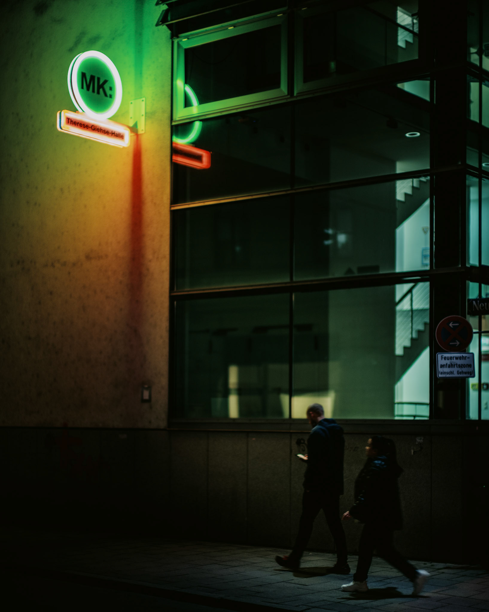 under the neon glow of munich's city center, captured with the exceptional leica noctilux f/0.95 mm lens, two figures traverse the quiet night. the green and red neon lights create a surreal ambiance, casting reflections on the glass facade and painting the scene with an urban mystique. this photograph encapsulates the calm of the nocturnal city, where modern architecture meets the solitude of the night, revealing the hidden whispers of the urban landscape.