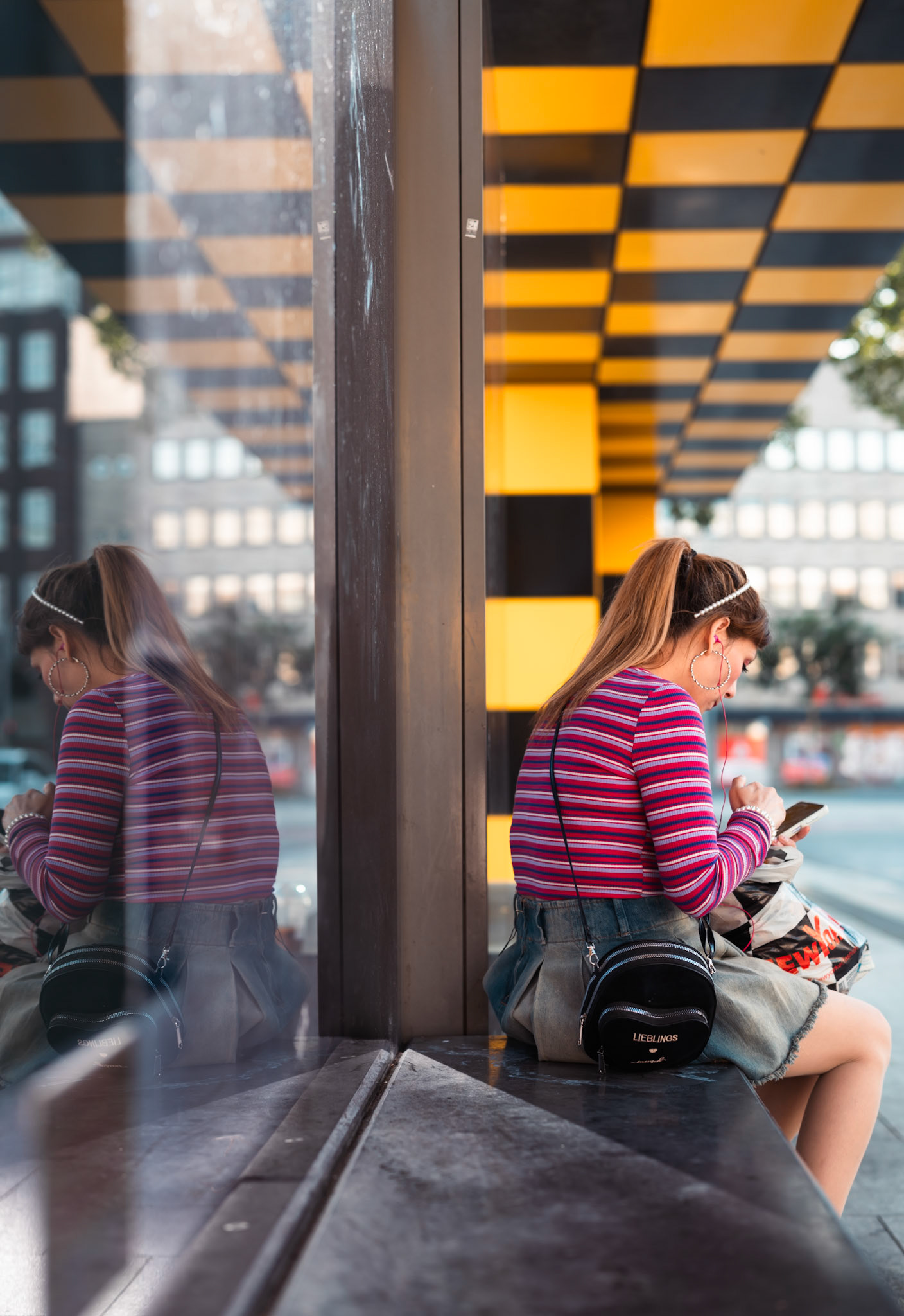 a candid moment captured at a city bus stop, where reflection meets reality. the scene plays with symmetry and color, as the bright yellow and black pattern frames the woman engrossed in her own world. the reflection in the glass creates a dual perspective, inviting the viewer to pause and ponder over the small stories unfolding in the everyday hustle. it's an intersection of urban life, color, and solitude, where even the quiet moments find their voice.
