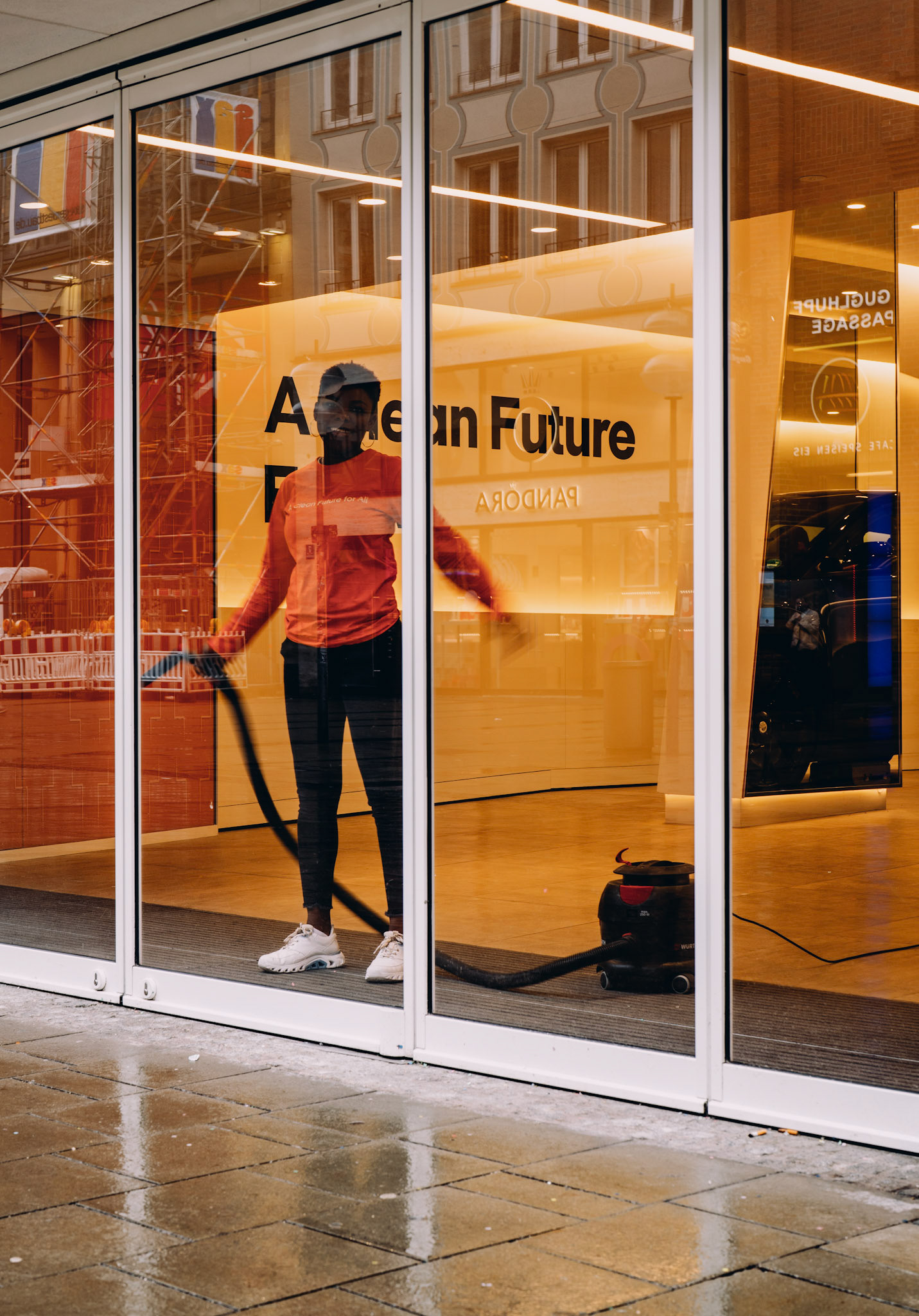 in a rainy munich, a cleaning lady in orange becomes part of the store's vibrant message of a clean future. amidst the drizzle, her silhouette is a burst of warmth against the coolness of the rain. she moves methodically, her presence as much a part of the storefront as the promise emblazoned above. the world outside continues its wet whisper, but inside, there's a bright insistence on order and optimism. even as the rain blurs the lines, her efforts carve out a space where clarity reigns.
