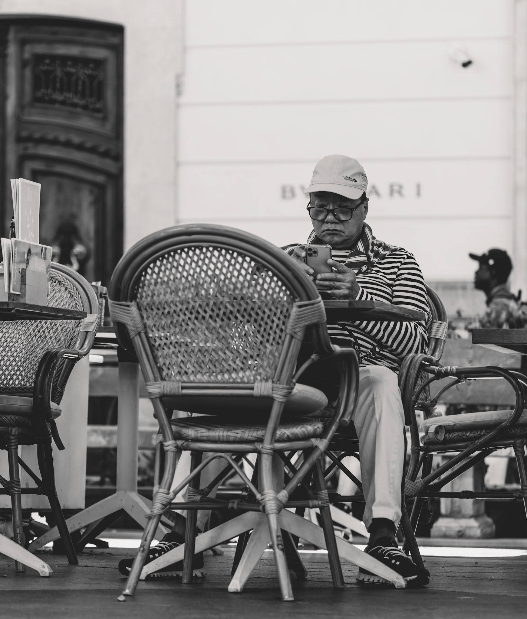 captured on the paseo del borne in palma, this black-and-white photograph reveals a man deeply engrossed in his smartphone, sitting amidst the classic charm of outdoor café chairs. his striped shirt and casual cap suggest a leisurely day, while the surrounding elements, including the iconic bvlgari sign and the textured architecture, provide a backdrop that blends history with the present. the image quietly contrasts the tranquility of a moment spent alone with the bustling life of the city around him, encapsulating a scene of modern contemplation.