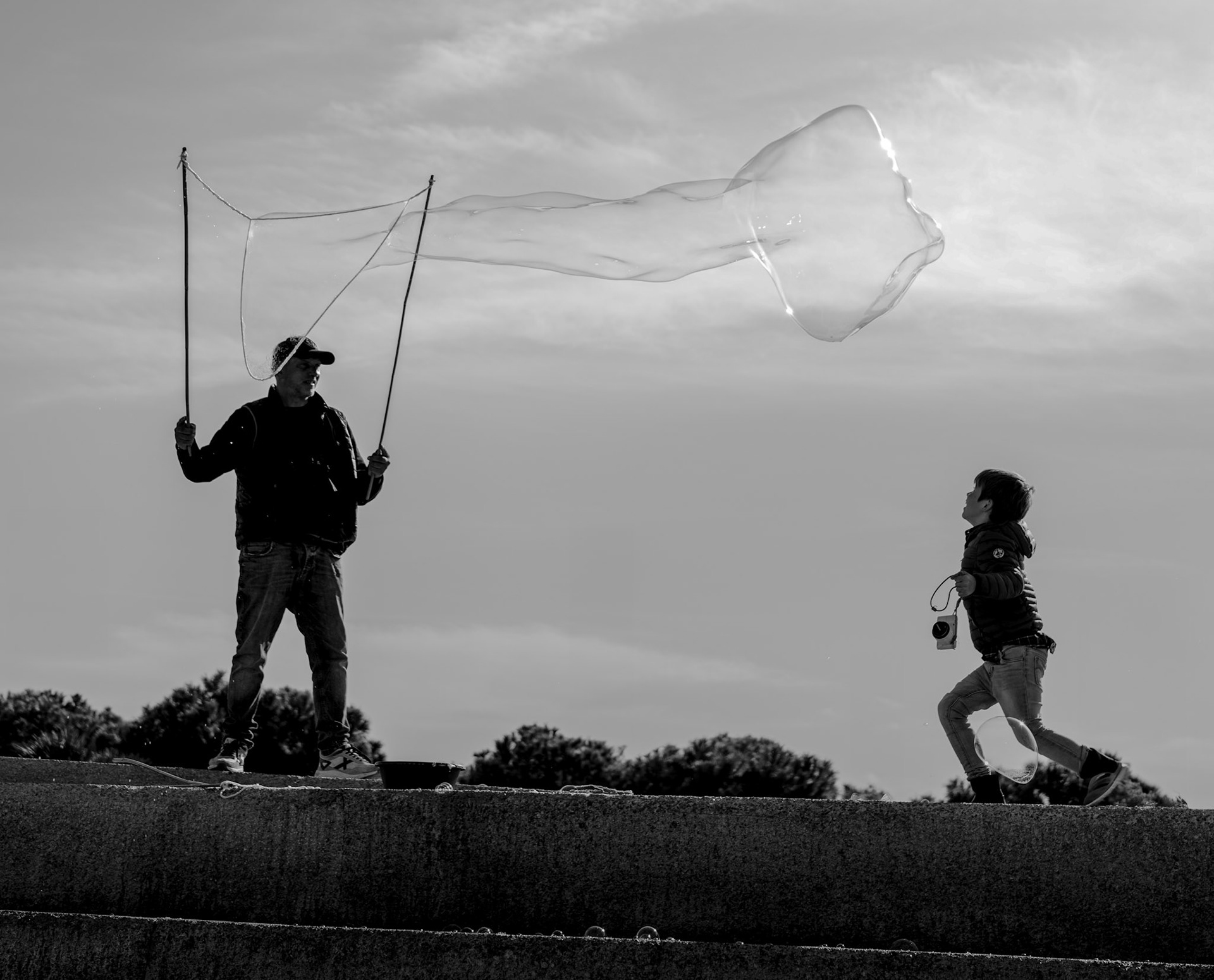 in palma's heart, by the parc de la mar, a child leaps towards the sky, chasing the graceful dance of a soap bubble, a fleeting giant conjured by the deft hands of an artisan of whimsy. it's a silent ballet against the canvas of a cloud-dappled sky, a moment where joy and wonder are as palpable as the sea breeze. the child's eyes are wide, feet barely touching the ground, suspended in mid-air like the bubble he pursues, and there's a sense of something magical at play, something of dreams and the pure, unbridled reach for them. in this dance, there is no before, no after, just the perfect now.