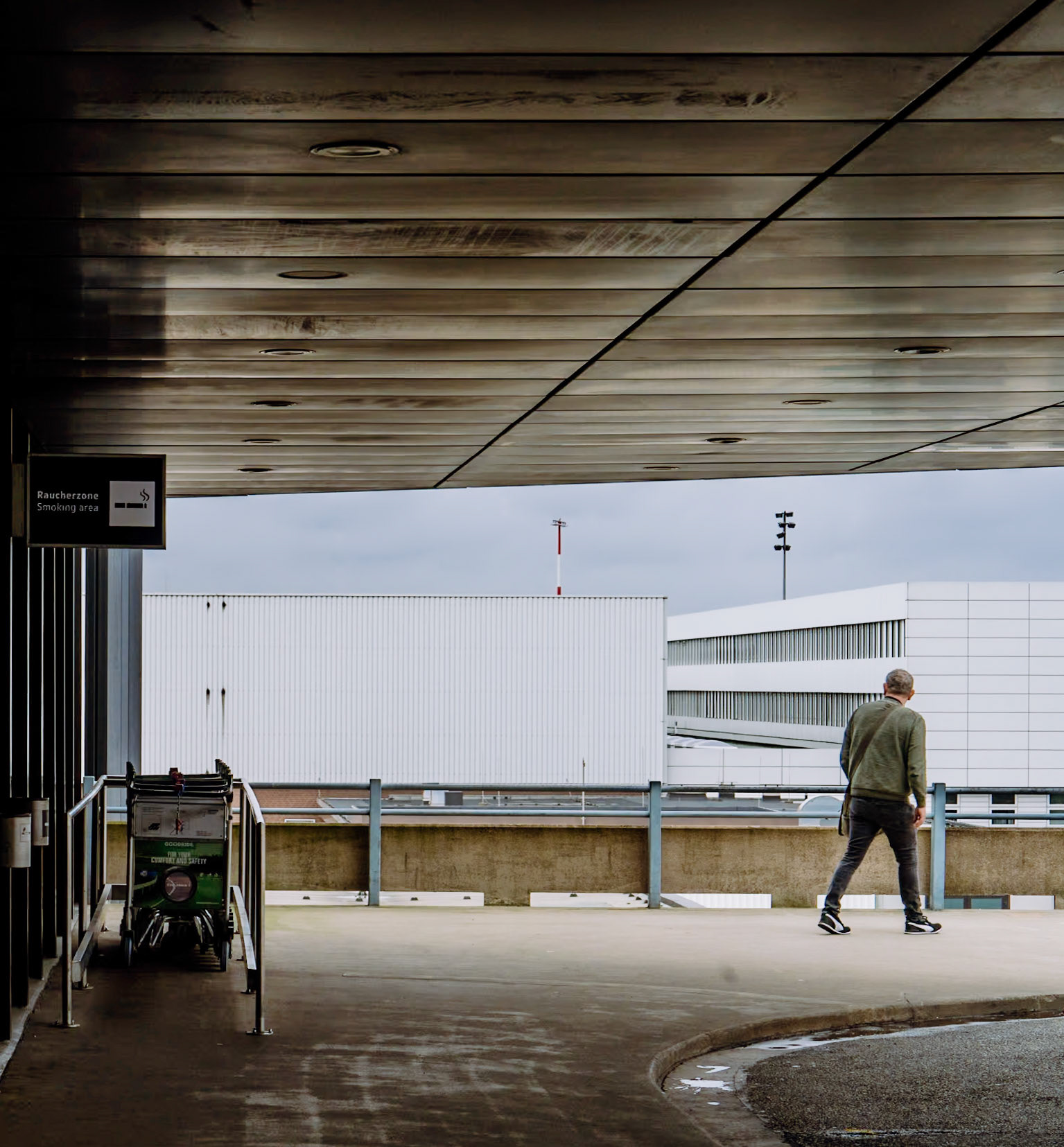 beneath the clinical gaze of fluorescent lights and the muted chatter of distant travelers, a lone figure traverses the urban expanse of hannover airport. each step echoes a subdued defiance against the rigidity of his surroundings. this is a dance of light and shadow, a fleeting moment captured where the vast, industrial structures meet the quiet solitude of a passerby. the dark overcast sky seems to weigh down, yet he moves unburdened, towards a horizon obscured by man-made giants. the rain has just left its gloss on the tarmac, reflecting not just the man but also the mood of this scene: introspective, a bit melancholic, yet undeniably hopeful.