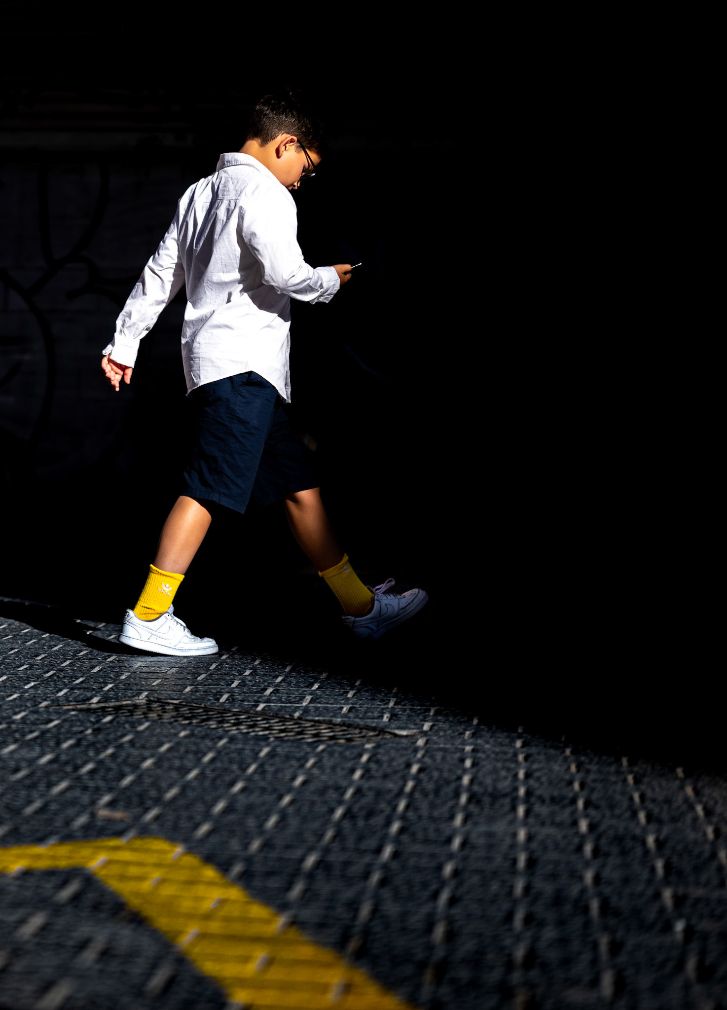 amidst the timeless charm of palma's old town, near the bustling Mercat de l'Olivar, a young boy strides purposefully, engrossed in his own world. the interplay of shadow and light frames his journey, casting an urban dance on the cobblestone street. his bright yellow socks and the hint of a yellow arrow on the pavement create a striking visual connection, a momentary burst of color in the darkened alley. this fleeting moment, where modernity meets tradition, captures the essence of a city in perpetual movement, where every step tells a story.