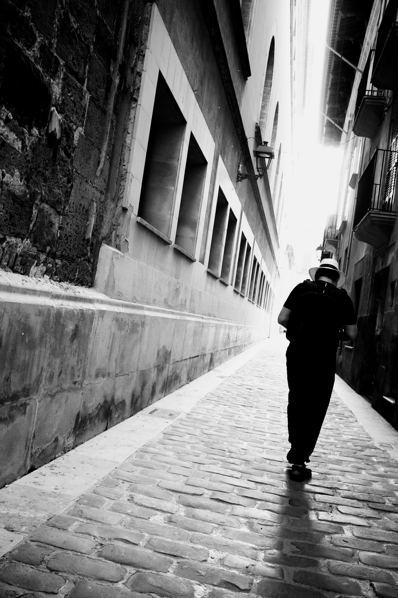 in the narrow alleyways of calatrava, the echoes of old palma whisper stories of yesteryear. a solitary figure, adorned in a straw hat, strolls through the cobblestone path, casting long shadows against the aged stone walls. the timeless feel of the black and white capture evokes a sense of nostalgia, where every step reverberates with the history of those who have walked these streets before. the man with the hat, a recurring motif in street photography, symbolizes an era of elegance and mystery, often serving as a bridge between the past and the present. in this instance, the straw hat of a modern tourist adds a touch of irony, blending homage with a subtle parody. it highlights the blend of old and new, tradition and tourism, paying tribute to iconic figures while gently mocking the clichés of contemporary travel. the play of light and shadow, the texture of the ancient bricks, and the lone silhouette create a poignant narrative of solitude and reflection in the heart of palma de mallorca.