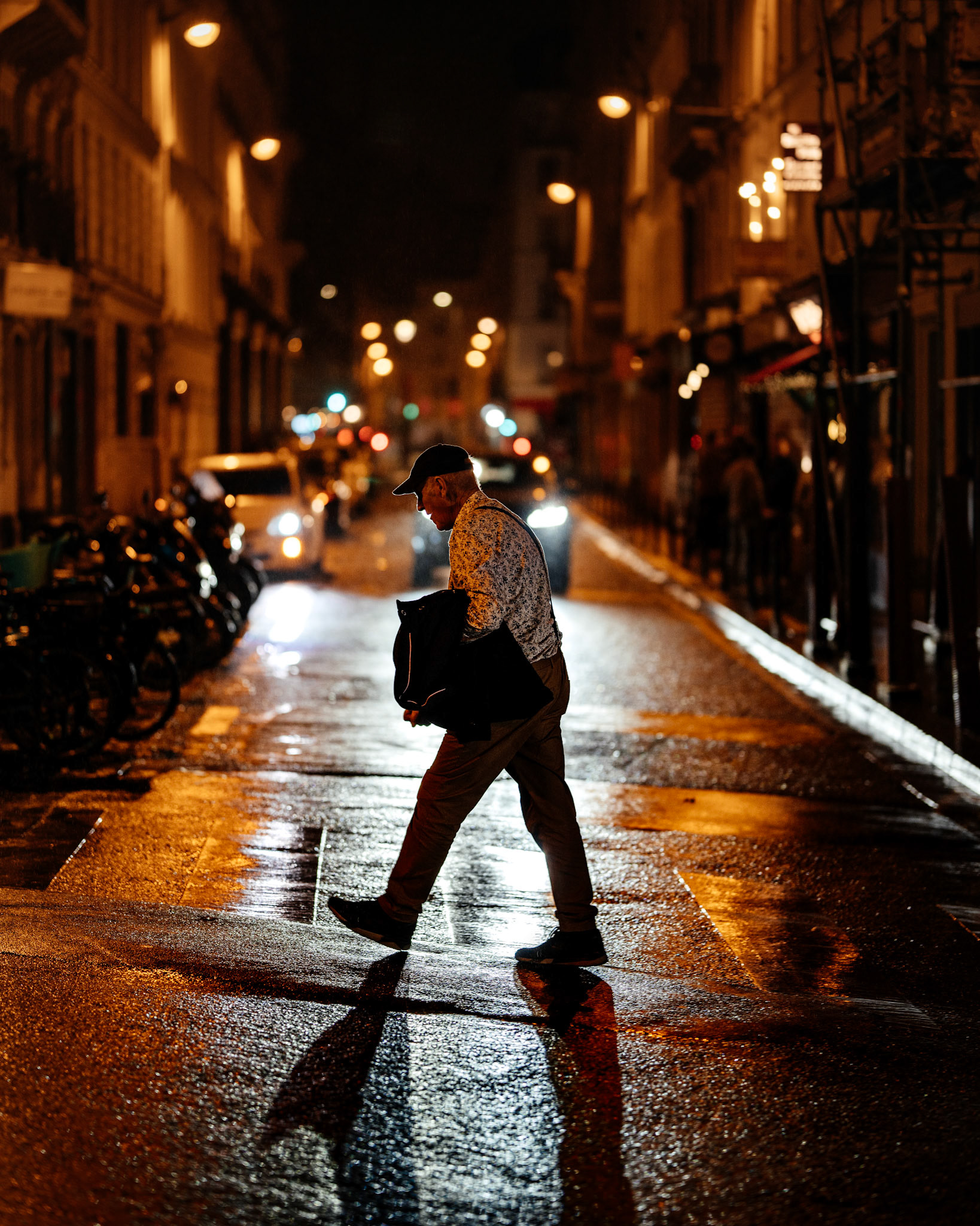 late at night, with rain-slicked streets reflecting the golden glow of streetlights, a lone figure crosses the empty lane. this moment of solitude amidst the city’s usual buzz invites the viewer to wonder—where has he been, and where is he going? captured in a fleeting second, the stillness of this scene reminds us of the quiet beauty in simple city moments.