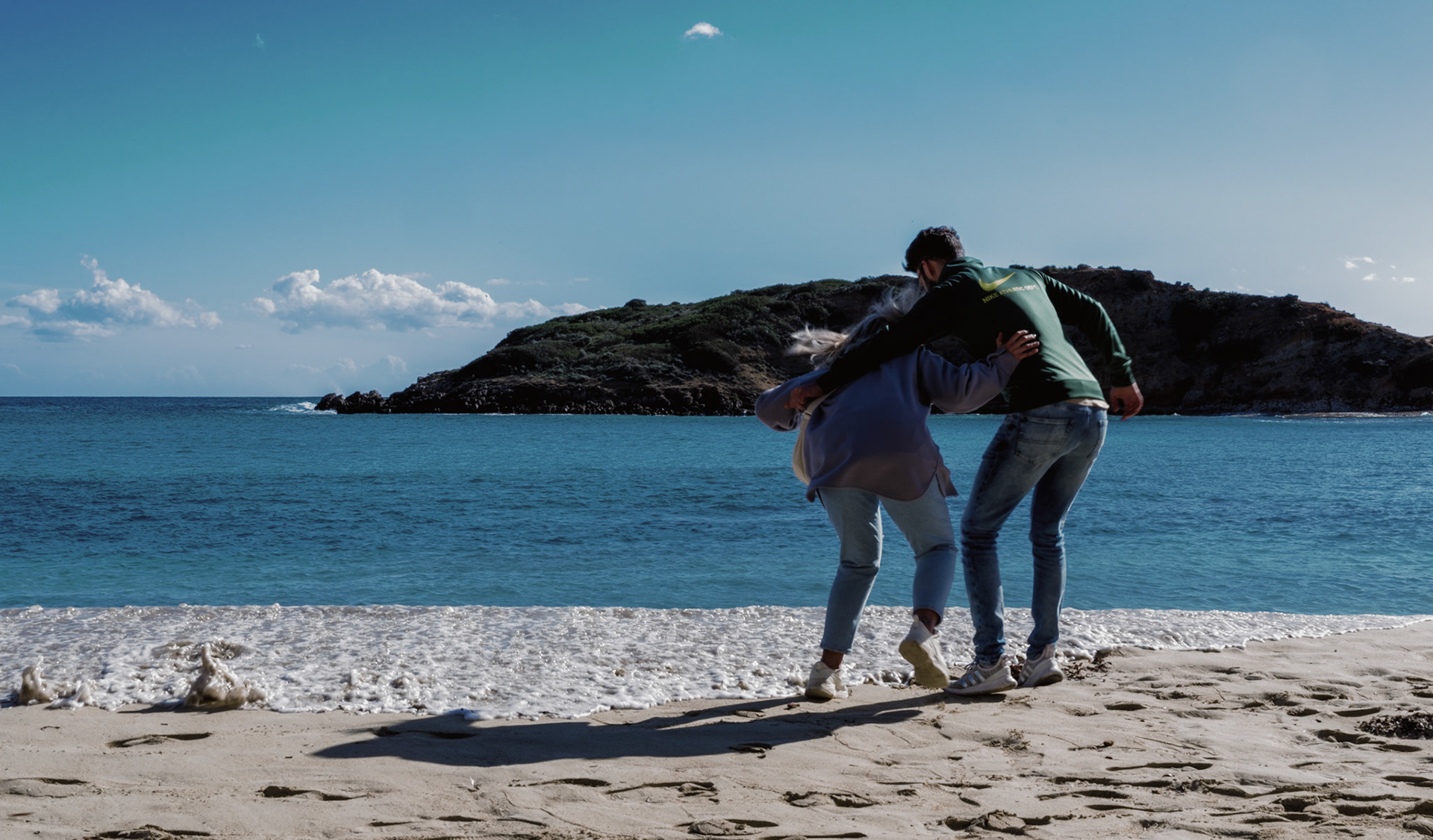 on the shores of puerto portals, a couple, wrapped in a cozy and intimate embrace, savor the view and each other's company. They are caught by surprise by the water, a playful reminder of the sea's spontaneous nature. The crystal blue of the water complements the clear sky, framing a moment where affection and the ocean's embrace meet, offering a snapshot of connection amidst the vastness of nature.