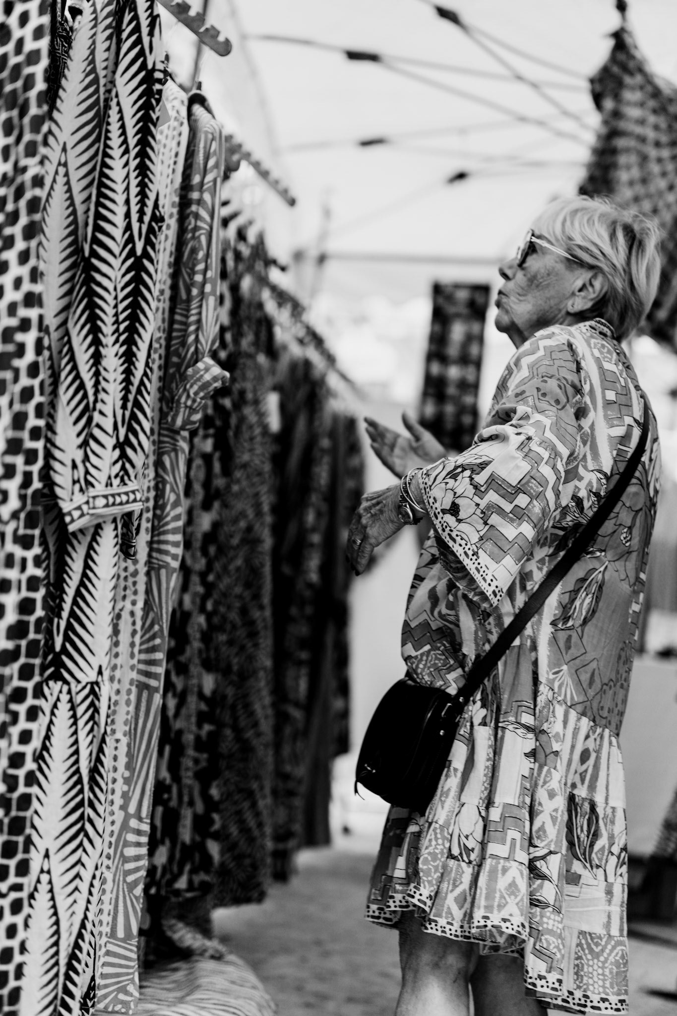a myriad of designs swirls around her as she browses the vibrant dresses on display. each pattern tells a story, each fabric a burst of creativity. her own outfit blends seamlessly into the cacophony of textures, creating a mesmerizing tapestry of shapes and hues. captured in the bustling saturday market of place des lices, this moment encapsulates the joyous chaos of colors and patterns that define the local sartorial landscape.