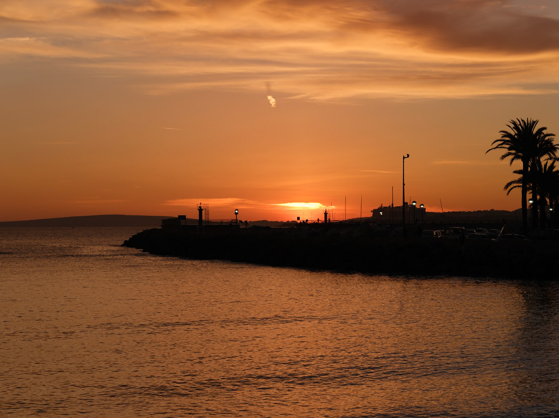 The sun dips low in Portixol, casting a fiery glow over the seaside district of Palma de Mallorca. The sunset is an ember in the sky, igniting the clouds and sea with warm hues. Silhouetted figures and palms are etched against the evening canvas, capturing a moment of tranquility at day's end. The gentle lapping of the water whispers farewell to the light, embracing the onset of twilight. It's a time when the bustle of the day yields to the calm of approaching night, leaving only the beauty of fading light and the promise of a starlit sky.