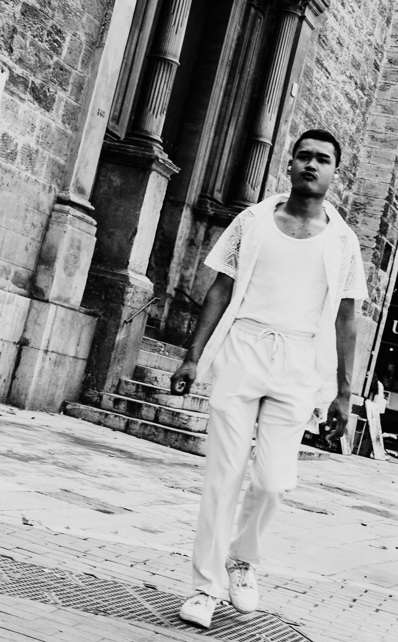 captured in the historic heart of palma de mallorca, this dynamic image showcases a young man confidently striding past the ancient stone walls and columns. the stark contrast between his modern, casual attire and the timeless architecture creates a striking juxtaposition, emphasizing the blend of old and new that defines the city. the diagonal composition adds a sense of movement and energy to the scene, making it a captivating street photograph.
