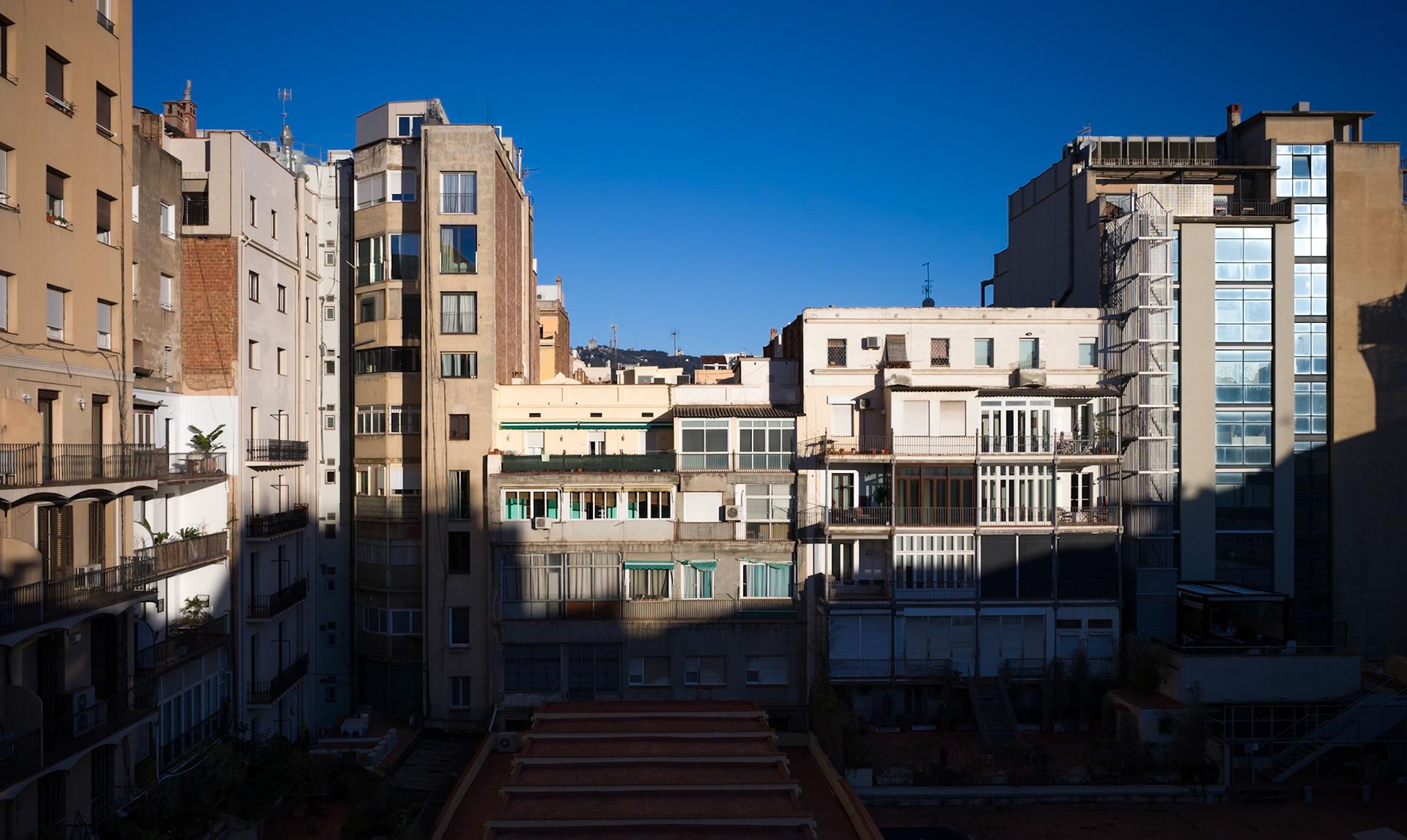 in the early morning light of barcelona, a hidden courtyard reveals the layered life of the city. buildings of different eras stand shoulder to shoulder, their facades a patchwork of history and daily life. the rising sun throws a sharp divide of light and shadow, carving out spaces where private lives unfold in the urban tapestry. balconies offer a glimpse into the quiet start of the day, as the city awakens in a hush, preparing to pulse with vibrancy once more.