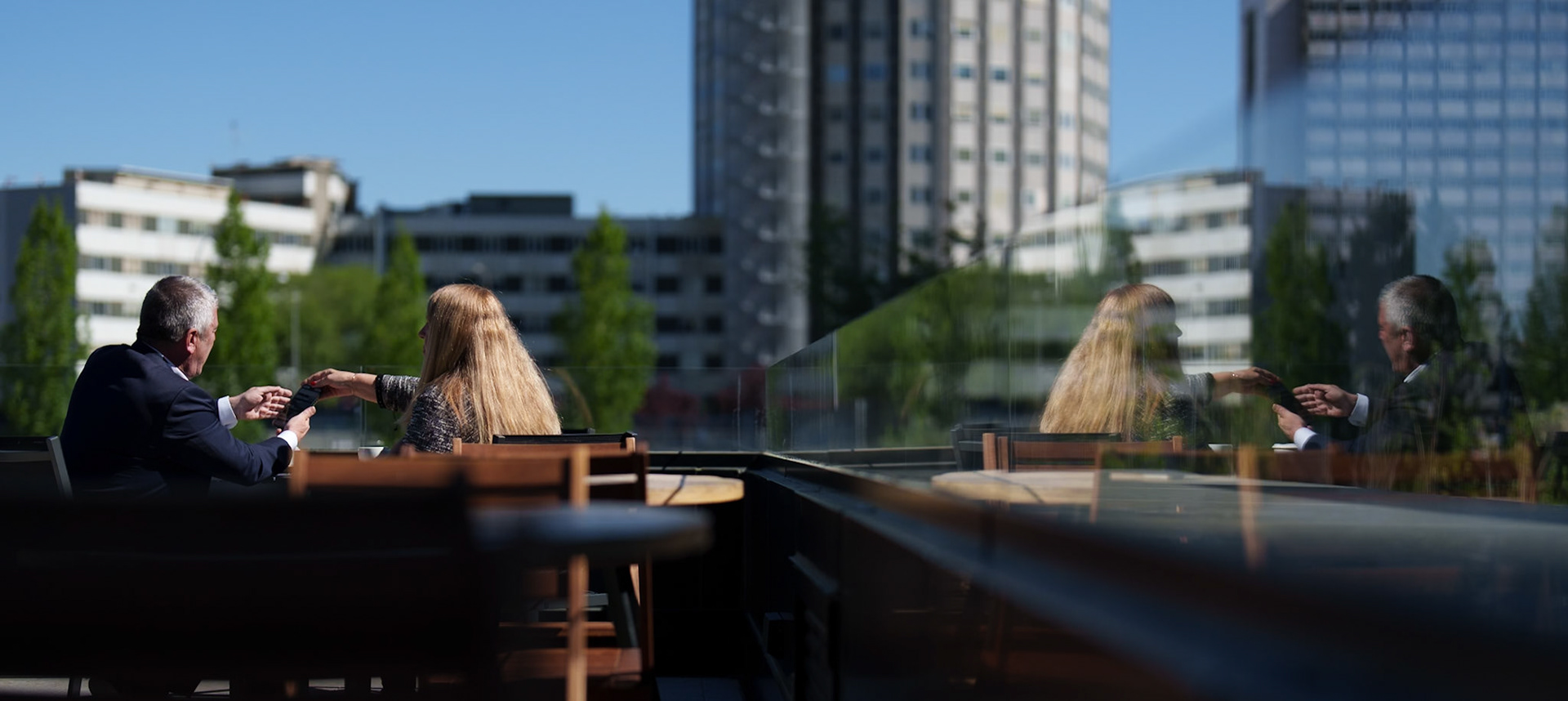 nestled amidst the towering testimonies of human ingenuity, two souls share a moment suspended in the clear, reflective dance of the present and the echoing past. seated in the gentle warmth of a madrid sun, their dialogue is more than mere words; it's an exchange of energies, mirrored not just in the glass that shields them but in the very essence of their interaction. the scene is drenched in contrasts—the soft organic lines of human figures against the harsh geometries of urban architecture—creating a tapestry woven with the threads of connection and reflection. here, the mundane act of sharing a drink transcends, reflecting the layers of personal and professional lives that intersect in the heart of the city.