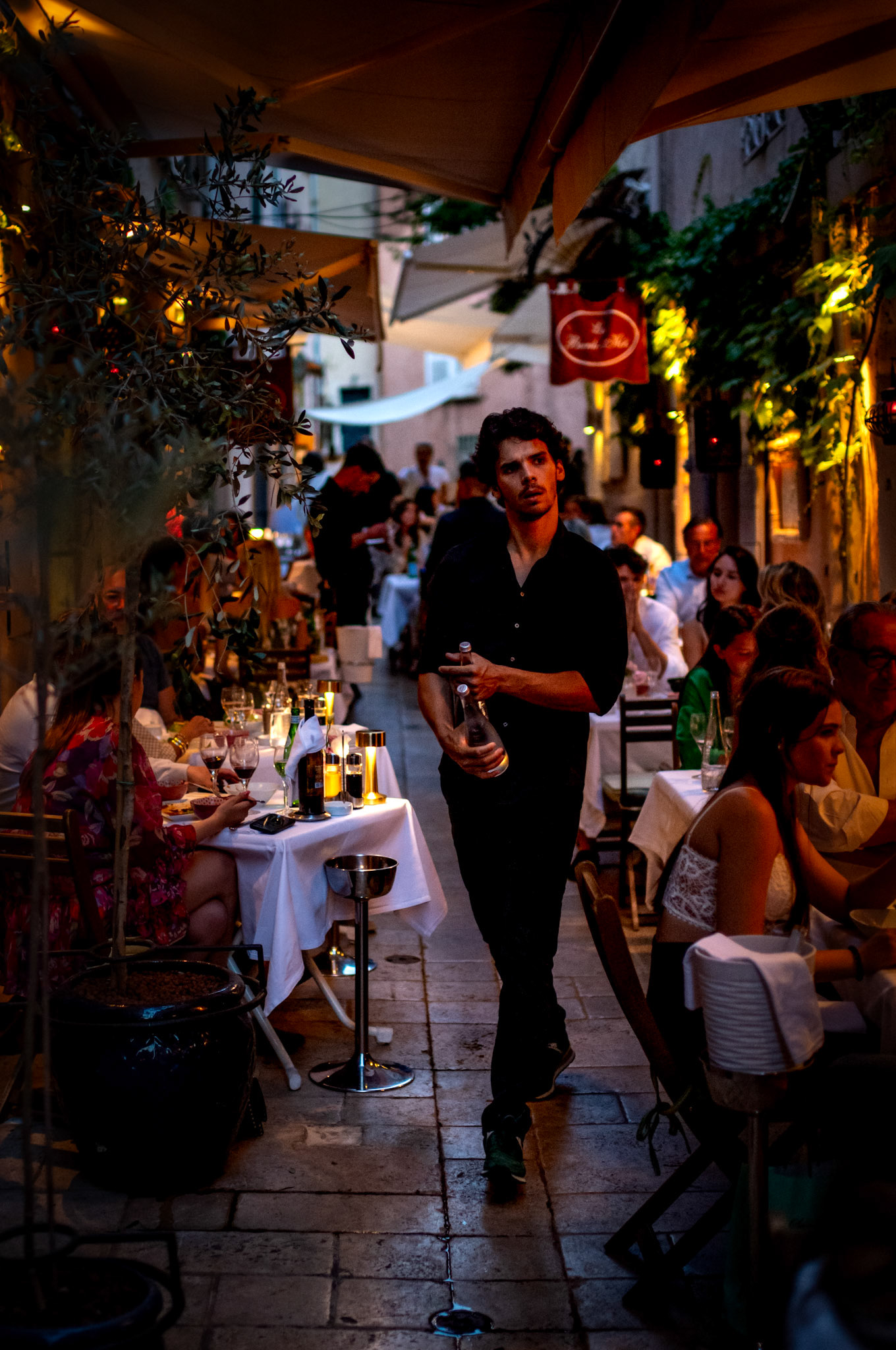 in the warm embrace of a mediterranean evening, nestled in a quaint alleyway of saint tropez, a waiter glides gracefully between the tables of ban hoi, an exquisite asian restaurant. the diners, bathed in the soft glow of candlelight, are lost in their conversations, savoring the delicate flavors of the night. the scene captures a moment of elegance and tranquility, where the atmosphere is as rich as the food, and every corner whispers a story.