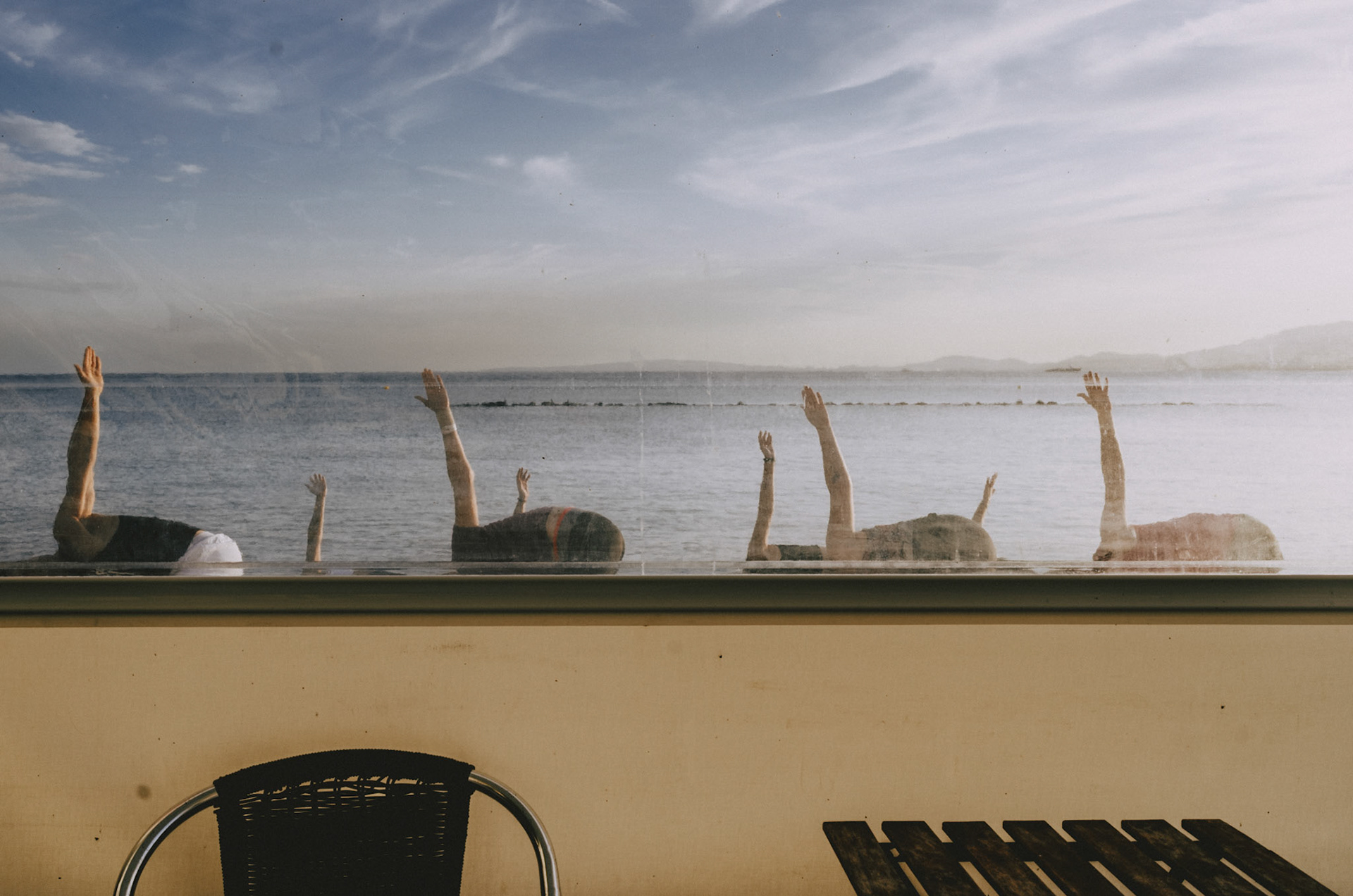 captured through the protective plastic sheet of the terrace at the charming old restaurant "el bungalow" in ciudad jardin, this image offers a serene yet surreal glimpse of a beach yoga class. the silhouettes of the participants, with their arms raised in various poses, blend harmoniously with the calm sea backdrop, creating a captivating dance of light and shadow. the juxtaposition of the outdoor scene with the empty tables and chairs of the restaurant adds a layer of quiet introspection, reminding us of the moments of stillness and mindfulness that exist amidst our daily routines.