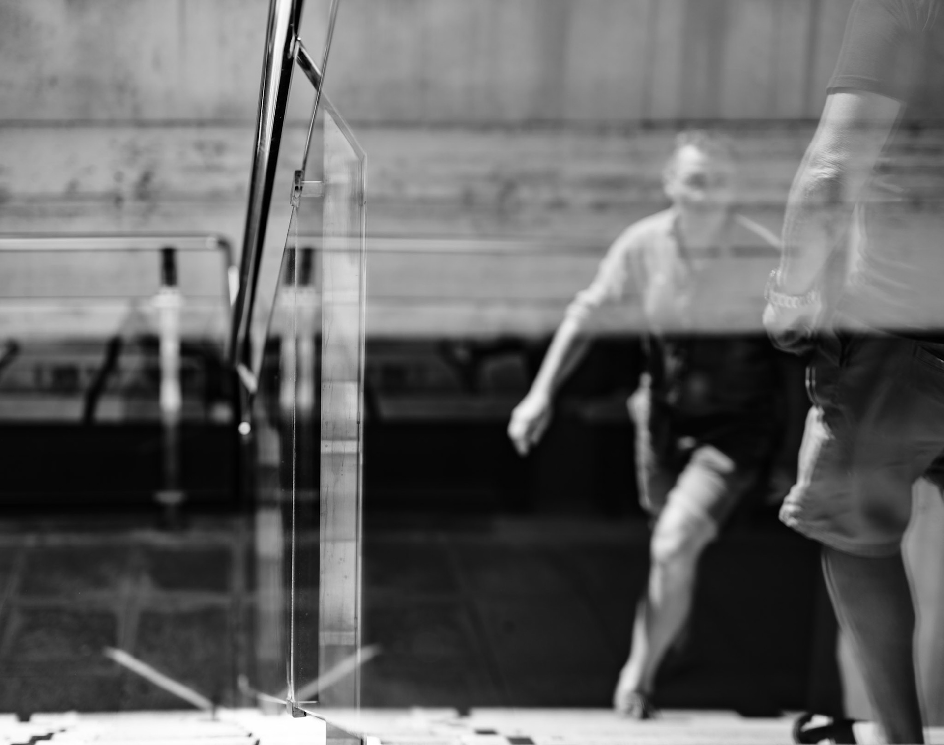 captured at the hospital of son espases near palma de mallorca, this image reveals a moment of fleeting movement. the blurred figures ascending the staircase, reflected in the glass, create a sense of transience and impermanence. the sharp lines of the railing contrast with the softness of the human forms, highlighting the intersection of structure and life. this black and white photograph encapsulates the quiet rhythm of everyday moments within the stark environment of the hospital.