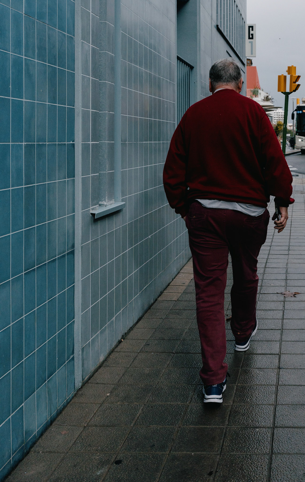 in palma's el terreno, a man walks in solitude, his red sweater a pop of color against the grey cityscape. the textured pavement underfoot and the tiled wall alongside him guide his steady stride. a simple moment of daily life, captured in the quiet before the streets awake.