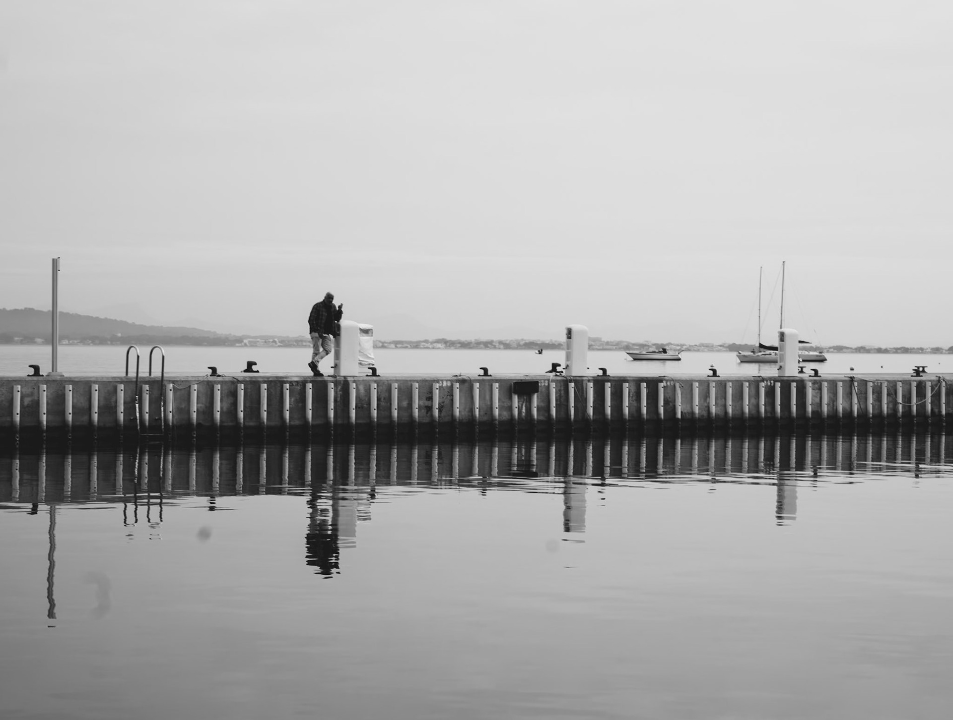 a serene black and white photo captures a moment on the pier with calm waters mirroring the scene. one man is gazing out over the dock where boats are moored in the distance. the stillness of the water creates a perfect reflection, enhancing the tranquility of the moment. captured in winter in port de pollenca.