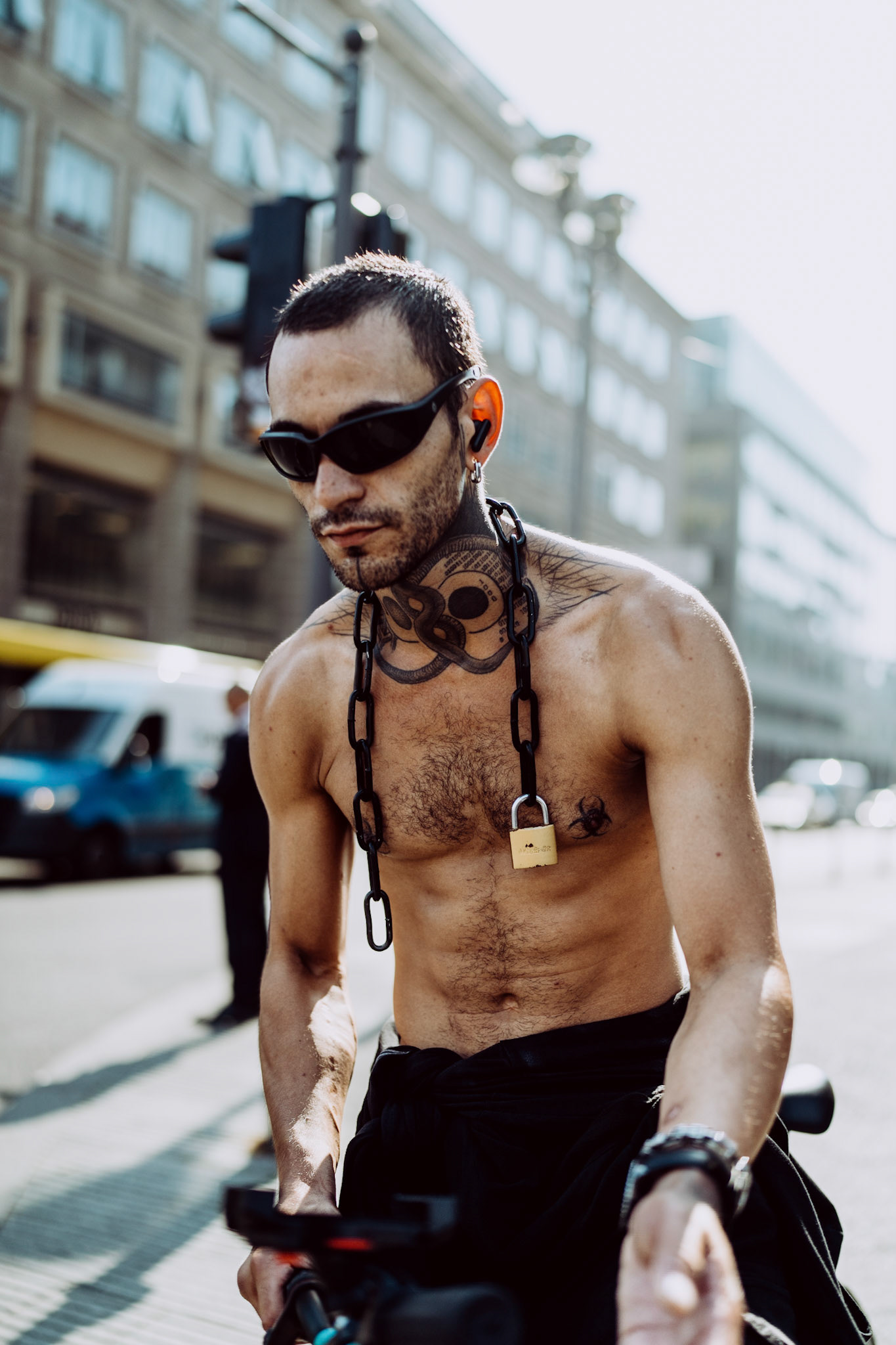 riding through berlin’s bustling streets, he embodies a mix of urban rebellion and street-smart confidence. the heavy chain with a padlock around his neck, contrasting his bare skin and dark sunglasses, speaks of the paradox between freedom and constraint. the sun cuts through the buildings of friedrichstraße, casting dramatic shadows, while the city moves in a rhythm only he seems detached from. it’s a moment of raw urban poetry, where style and intensity intersect.