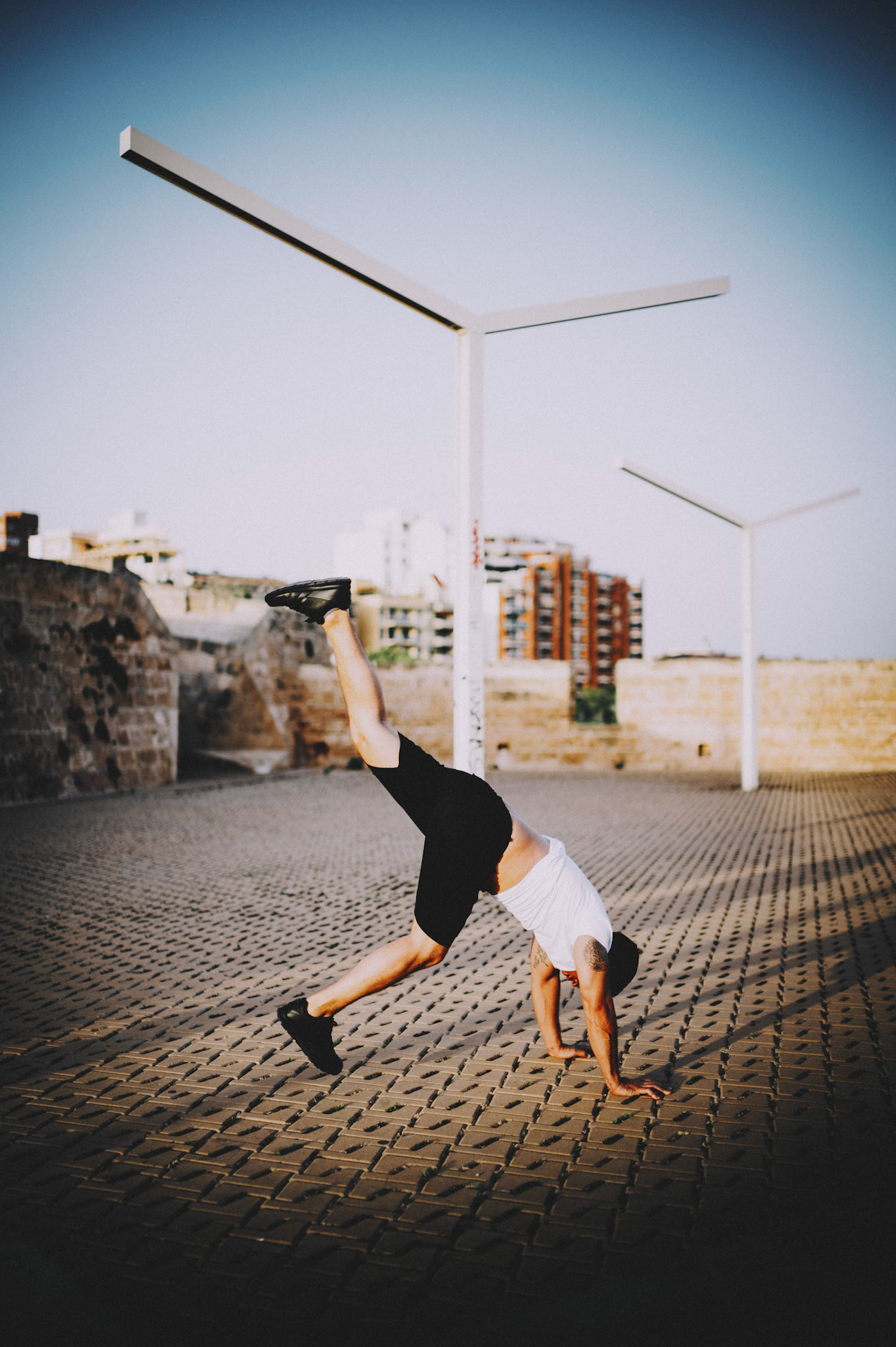 captured on the ancient city walls of calatrava in palma de mallorca, this moment fuses human strength with architectural elegance. as the sun begins its descent, the urban landscape stands still while the figure in motion defies gravity, balancing on hands with perfect poise. the geometric patterns of the pavement and the stark, modern lines of the streetlights juxtapose against the historical backdrop, creating a serene yet dynamic scene. it’s a dance of shadows and light, where every element plays a part in this symphony of tranquility and tension.