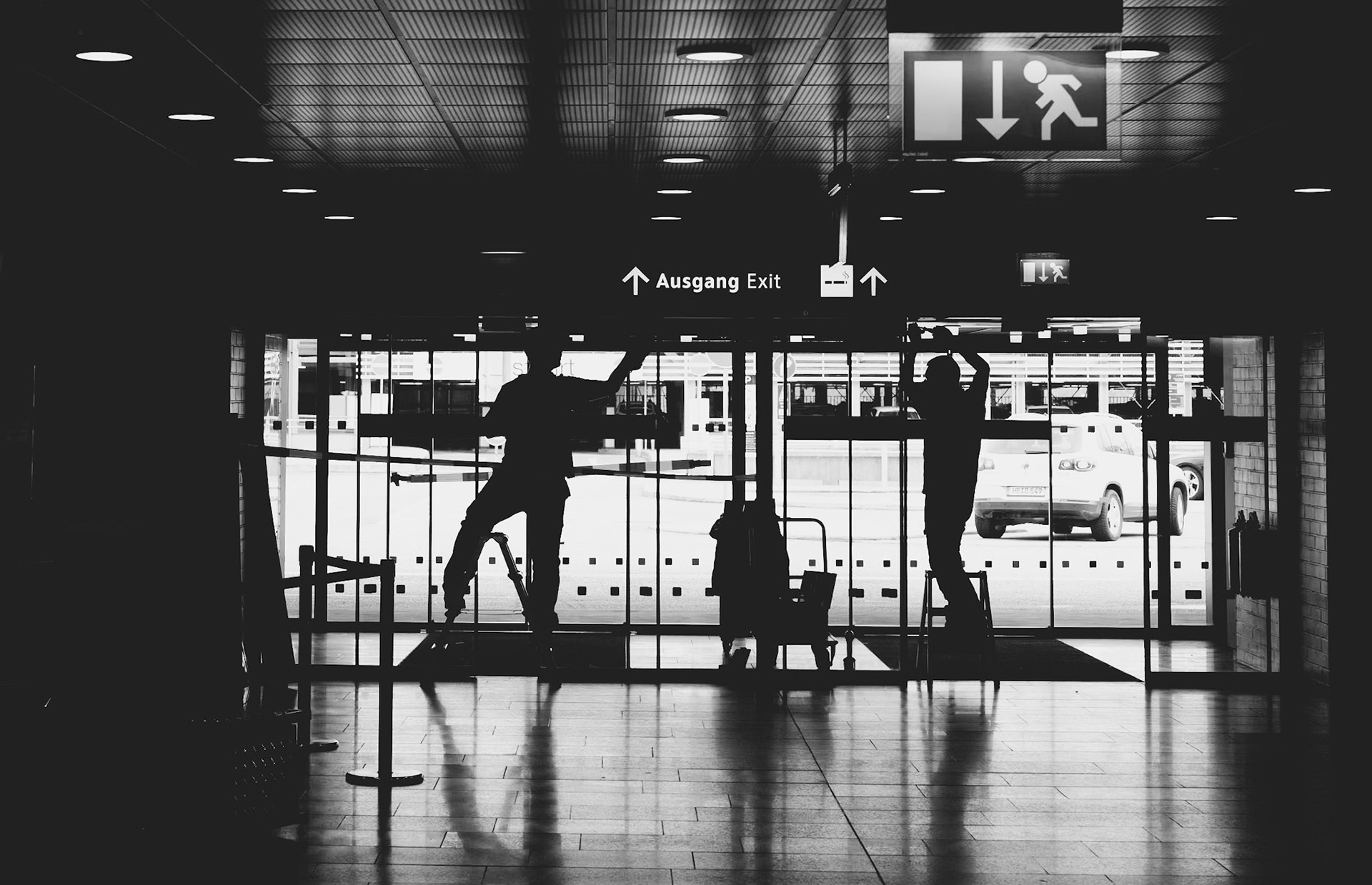 at hannover airport, a pair of maintenance employees engage in repair work on the automatic doors, a crucial task ensuring smooth passage for travelers. their silhouettes are framed by the doorway, tools in hand, against the backdrop of a busy terminal outside. this moment captures the essential yet often unseen efforts that maintain the airport's rhythm. their focus and coordination reflect the dedication behind the scenes, keeping the flow of movement uninterrupted and safe for everyone passing through.