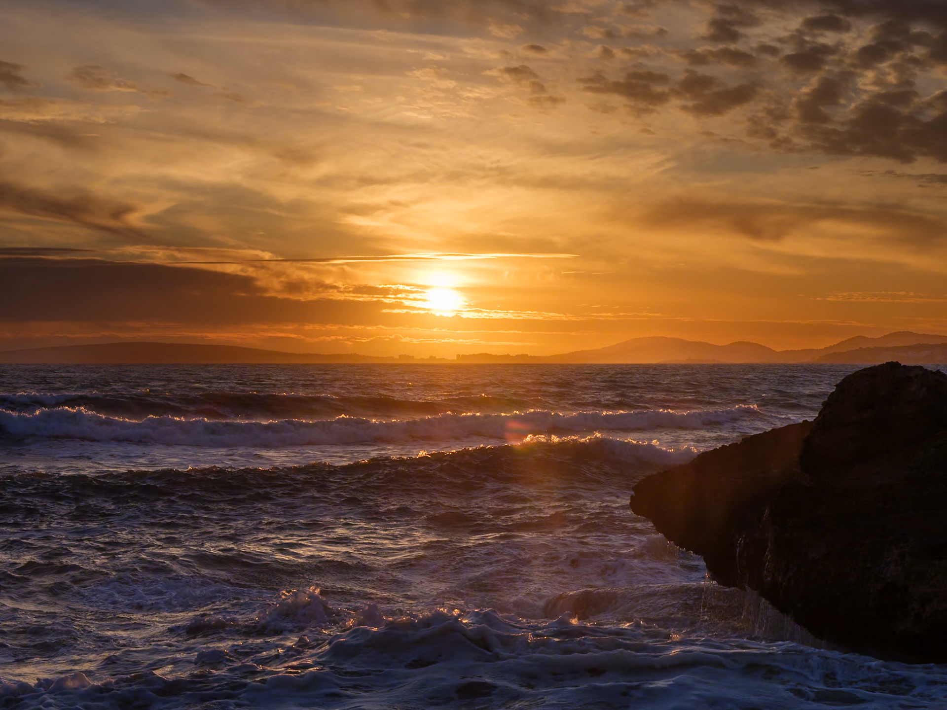 as the day's fiery orb descends, it kisses the sea, setting it ablaze with the last warmth of sunlight. waves, wild and untamed, rush towards the shore, eager to absorb the scattered embers of the fading sun. clouds, painted in strokes of apricot and mauve, drift languidly across the canvas of the sky. a solitary rock, steadfast and enduring, meets the ocean's embrace, a silent witness to the cyclical whisper of time. this is the dance of day and night, a performance of light on water, a spectacle that speaks to the soul's yearning for natural beauty and the quiet promise of the twilight hour.