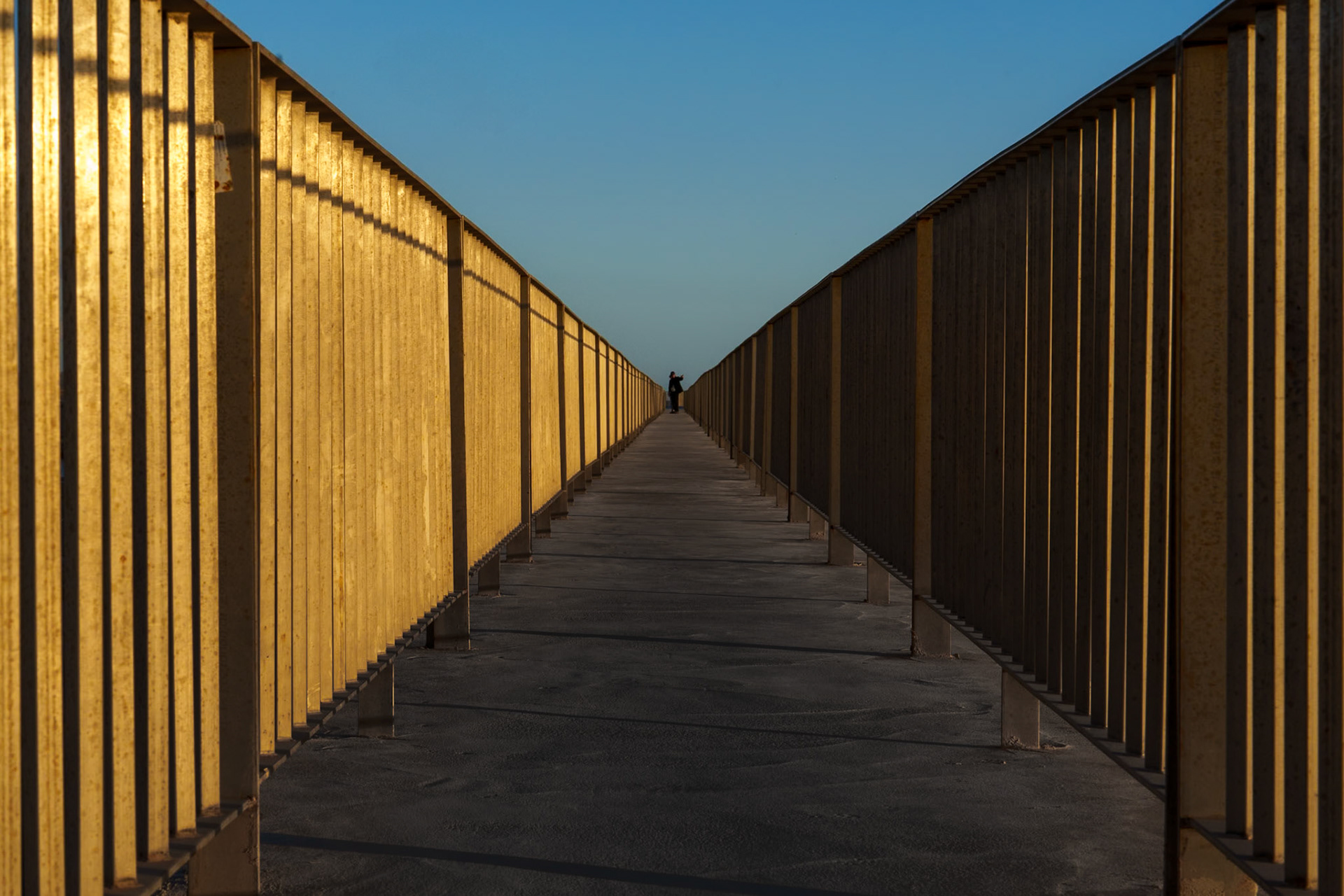 as the sun teeters on the brink of the horizon, its golden hues breathe magic into the iron lattice of a pathway in portixol's modest harbor. this isn't just a walkway; it's a corridor of time, capturing the essence of transition as day gives a slow nod to night. there's a solitary figure in the distance, perhaps a wanderer pondering the poetry of dusk, or maybe a local, steeped in the quietude of their regular passage. this place, where the sea's whispers meet the firmness of the land, becomes a canvas, the iron glowing like molten gold, reshaping a utilitarian structure into a spectacle of light and shadows. here is a moment of stillness, yet brimming with stories untold.