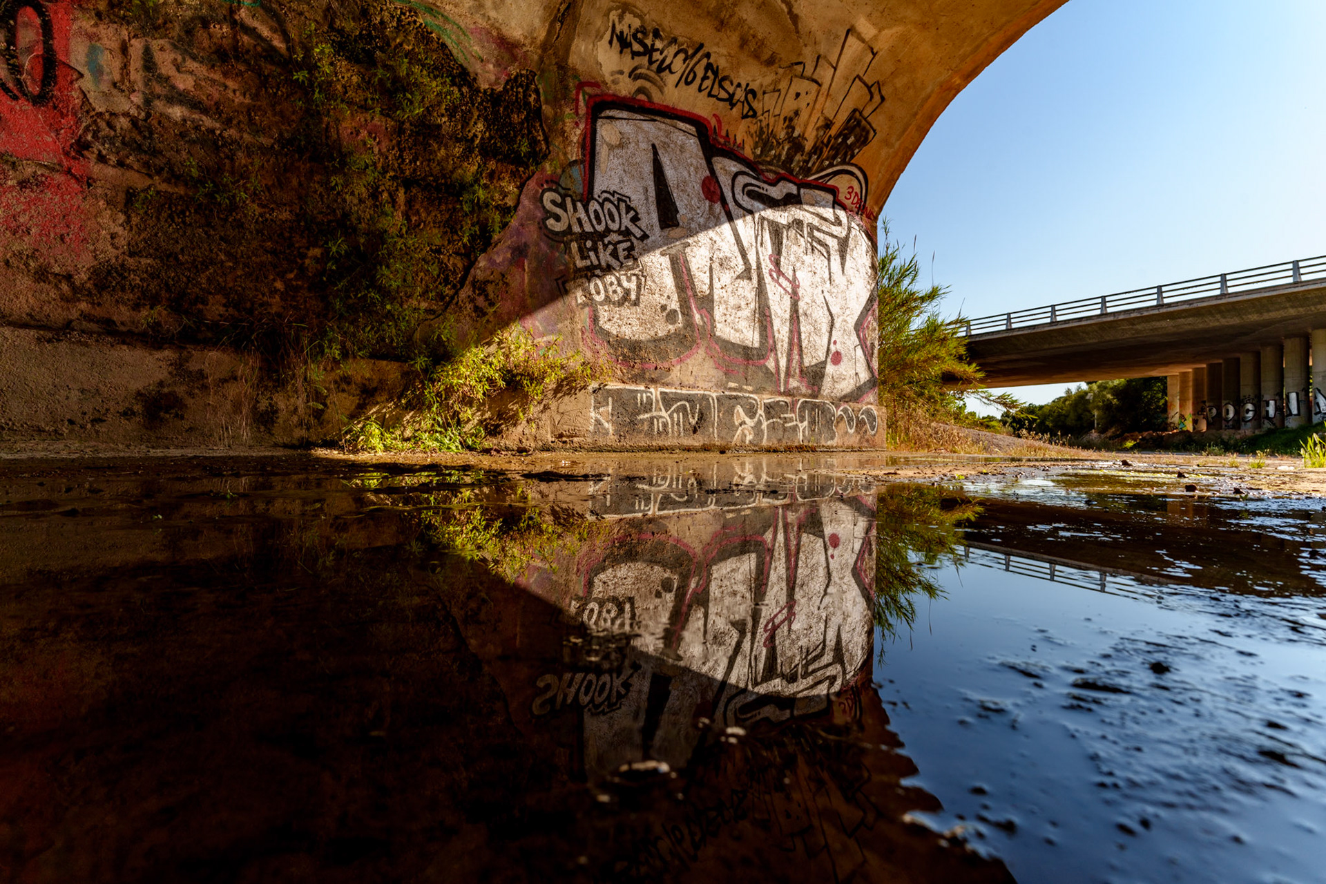 captured during a cycling tour in the hinterland behind santa ponça, this photograph showcases a striking reflection under a highway bridge. this green zone, perfect for hiking or mountain biking, culminates just before the town with this unexpected urban scene. water from various small stream beds uphill converges here, allowing for this stunning reflection in the puddle despite the bright sunshine. the contrast between the natural greenery and the graffiti art on the bridge’s concrete pillars creates a visually compelling image.
