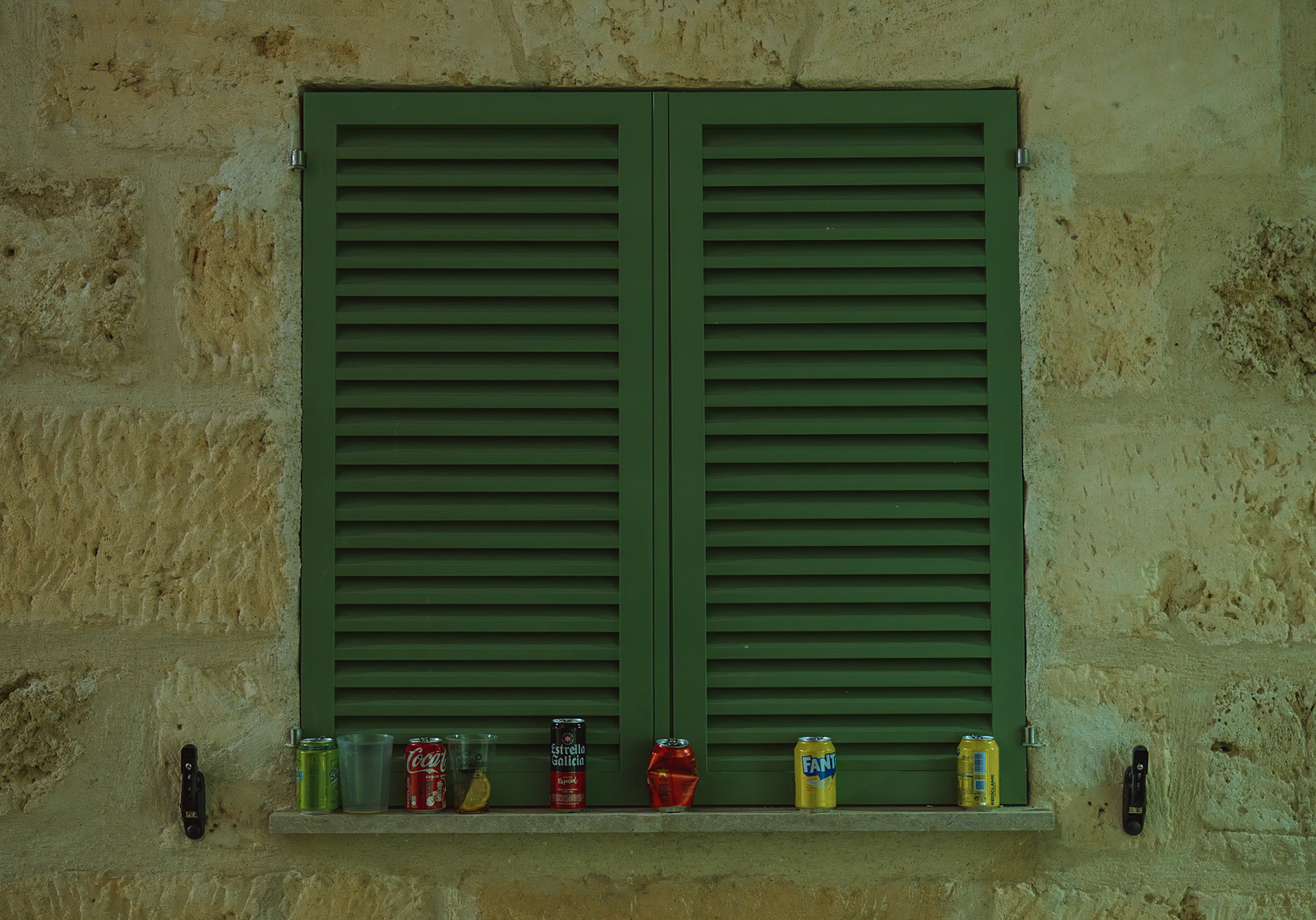 In the quiet rural village of sa pobla in mallorca, a stone wall with a green window shutter forms the backdrop for a simple yet evocative scene. lined up on the ledge are various cans of drinks, a quiet witness to the festive celebrations of san antoni, captured under the cloak of night.