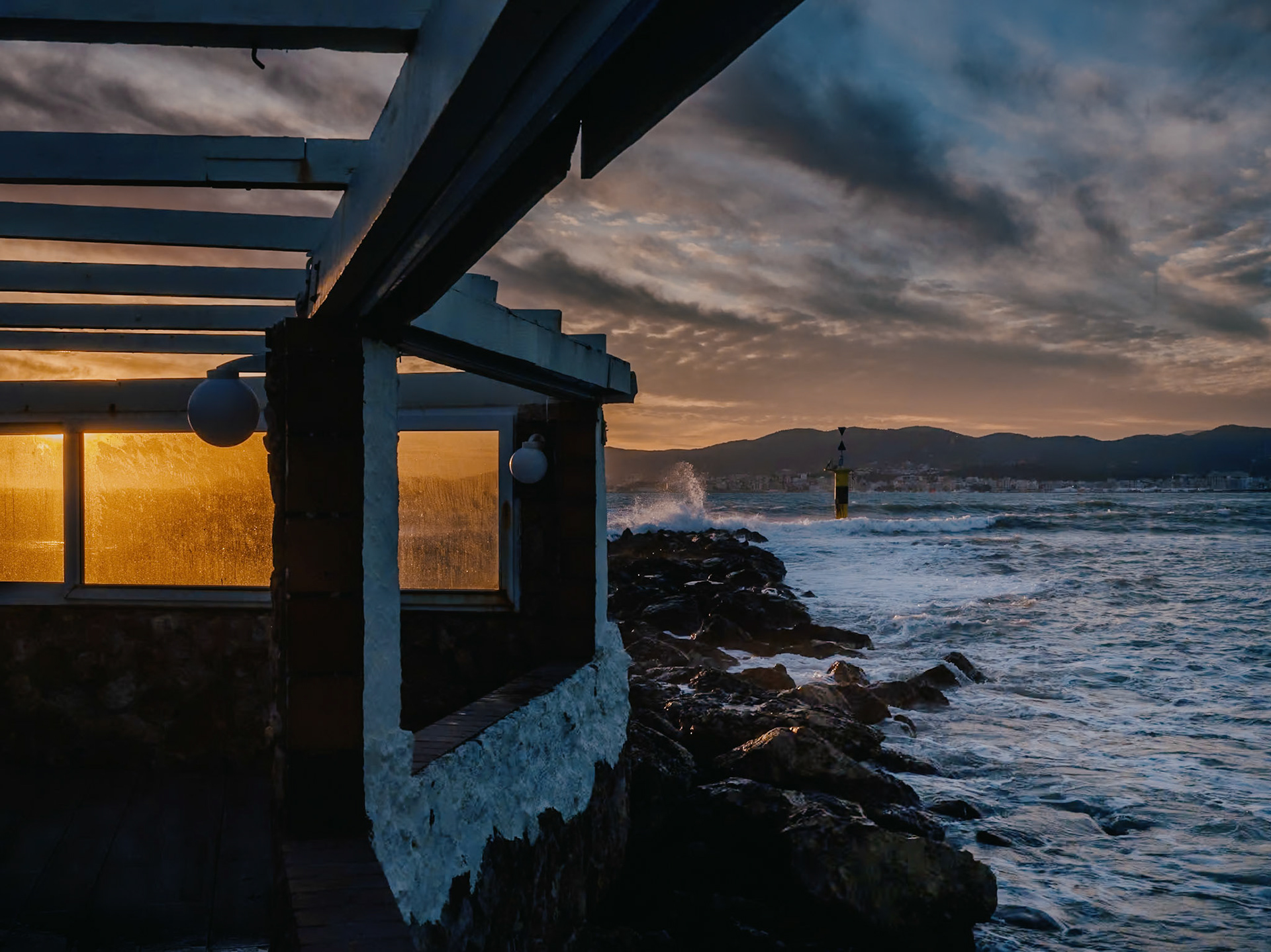 in the vibrant hues of winter's embrace, the terrace of el peñón 1957 becomes a stark contrast against the fervor of a storm. as the sun sets, casting a golden glow over palma de mallorca, the waves clash against the shore with a rhythmic persistence. the silhouette of the restaurant, a steadfast presence, reflects the sun's fleeting moments, while shadows dance upon the walls, marrying the chaos of the storm with the peace of the ending day.