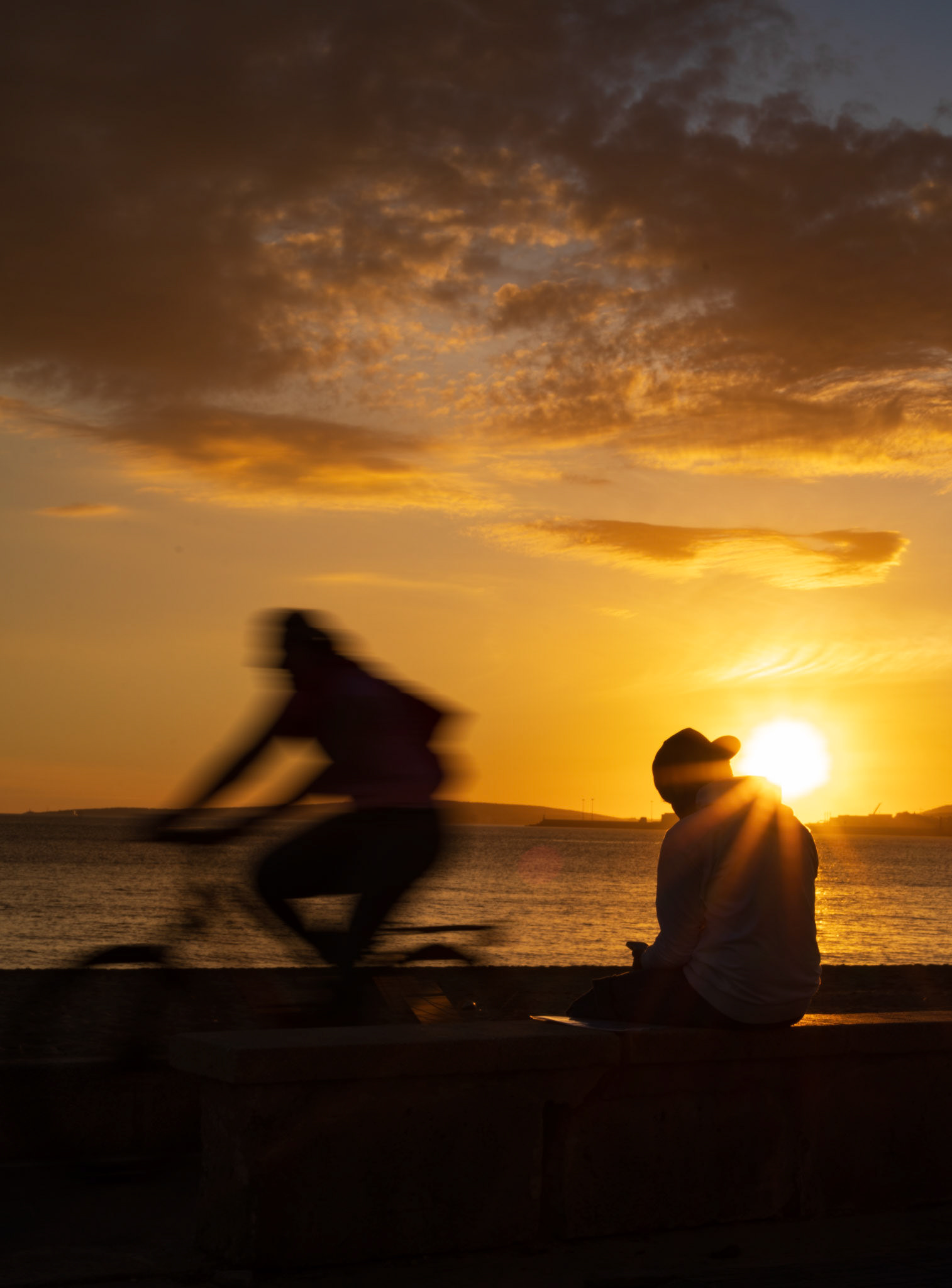 this photo captures a serene and fleeting moment by the sea in portixol, a district of palma de mallorca, just as the sun dips below the horizon. the sky, bathed in golden hues, echoes the warmth of the day slipping away. in the foreground, a cyclist moves swiftly through the frame, a blur of motion against the calm, still silhouette of a person absorbed in thought, seated on the promenade. the juxtaposition of the blurred cyclist and the stationary figure speaks to the passage of time and the quiet reflection that often accompanies the end of a day. the scene encapsulates the ever-changing rhythm of life in this coastal town, where the day’s end brings a moment of stillness and contemplation.