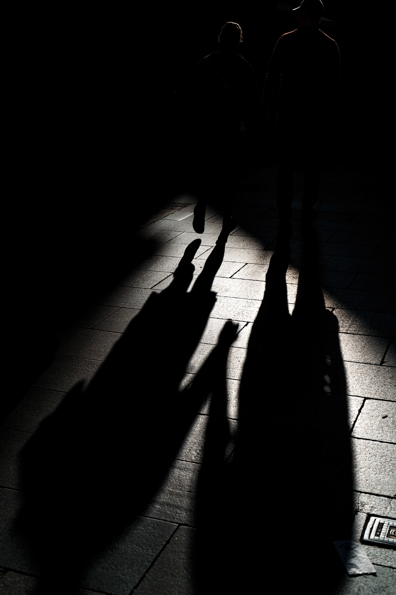 two figures walk hand in hand through a narrow corridor of light, their forms reduced to shadow and silhouette. the faint outline of their linked hands adds an unexpected warmth to the scene, turning an ordinary walk into a moment of quiet intimacy. with faces hidden and only their shadows stretching long across the pavement, this image captures a fleeting but tender connection, a small gesture made mysterious in the play of light and darkness. it invites the viewer to imagine who they are and what stories they carry in this quiet, cinematic scene.