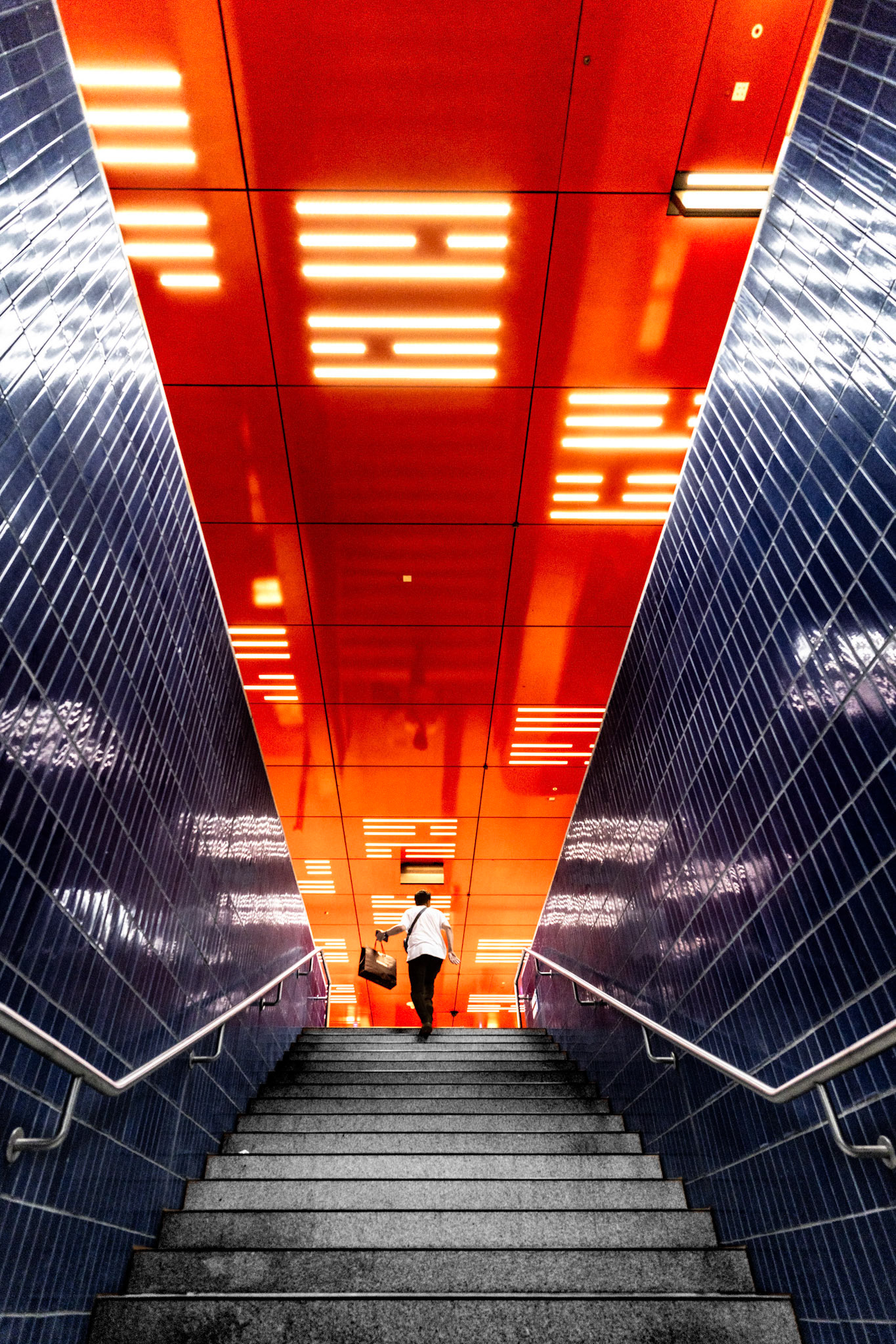 beneath the city’s surface, this moment captures the interplay of light and architecture, transforming an ordinary subway station into a vibrant canvas of color. the bold, fiery orange ceiling contrasts sharply with the deep blue tiles, guiding the eye upwards as if towards an urban horizon. the lone figure, mid-step, seems almost engulfed in this spectrum, their journey bathed in the glow of fluorescent patterns above. it’s a fleeting encounter with modernity, where every step is a rhythm in the symphony of city life, resonating with the energy of marienplatz in hannover, a place where routine meets the extraordinary.