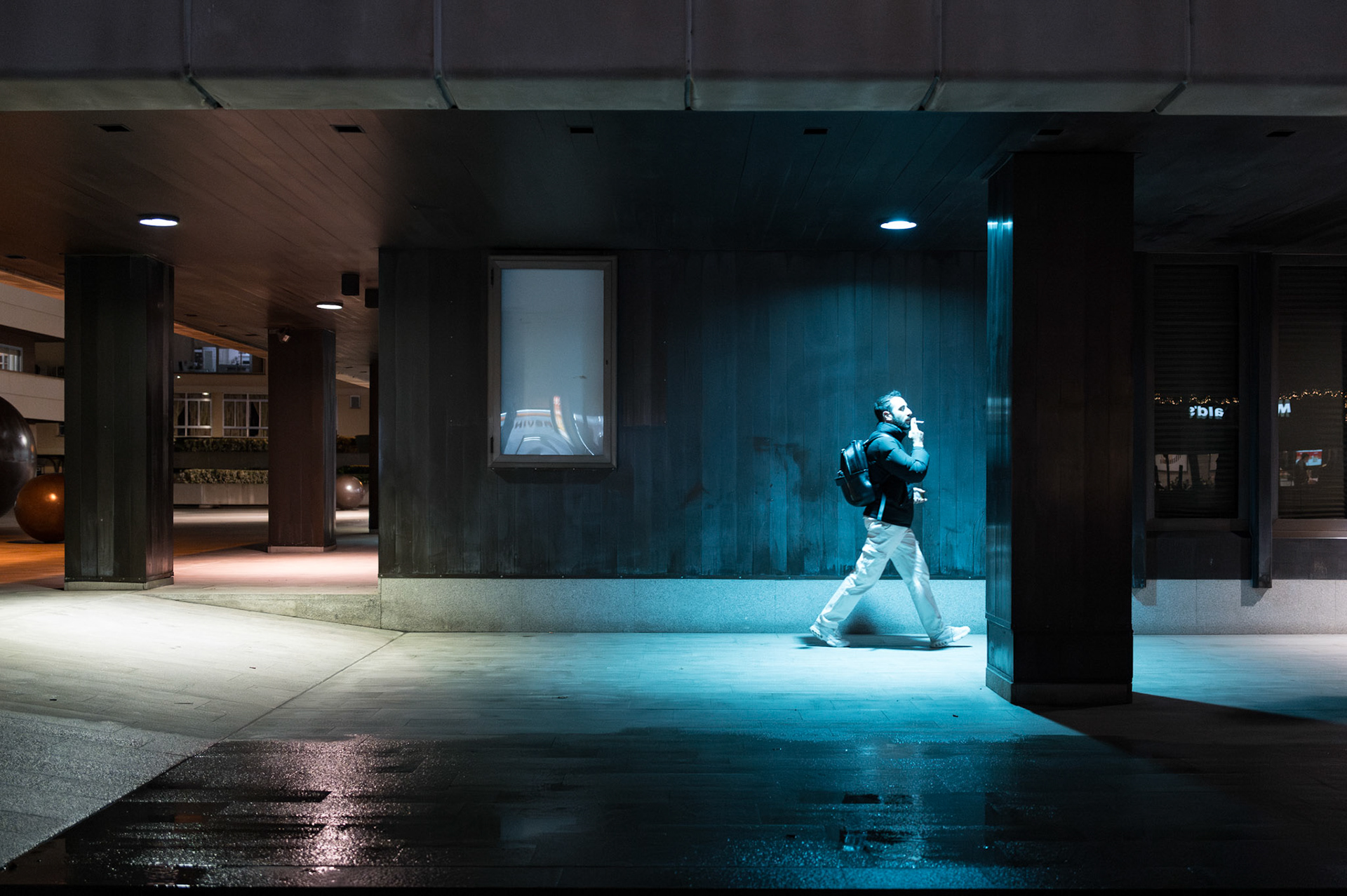 in palma de mallorcas district pere garau i captured this photo. under the neon glow, a man strides through the night. his posture relaxed, enjoying his cigarette, a backpack over his shoulder, he's illuminated by a striking blue light that contrasts sharply with the surrounding darkness. the city's pulse is felt in this solitary moment, a snapshot of urban solitude.