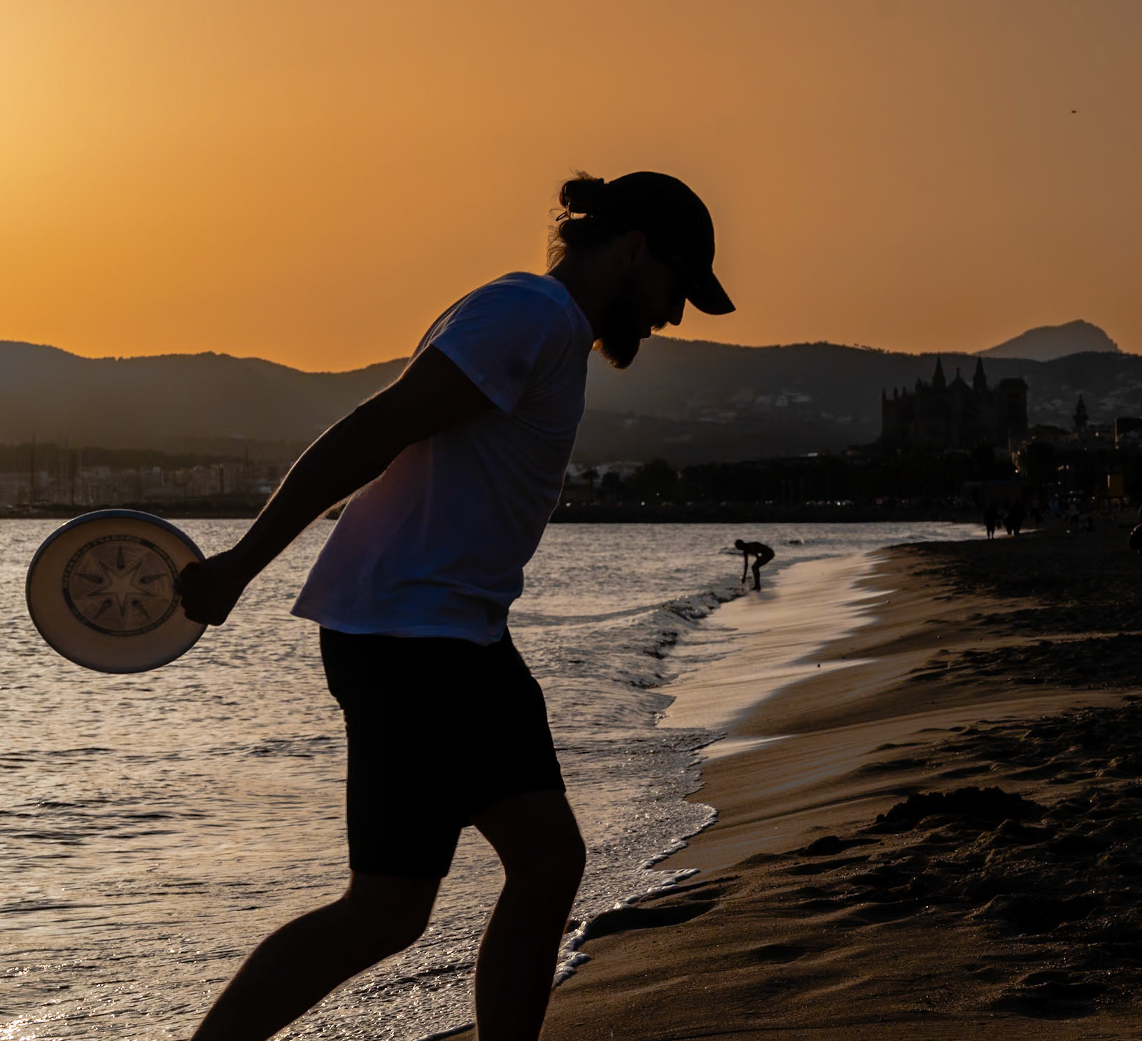 on the city beach of palma, as the sun dips below the horizon, a silhouette against the golden canvas captures the spirit of twilight. the iconic cathedral looms in the soft focus of the background, its spires gently blurred into the mountainscape, a quiet guardian over the evening’s serene play. a figure poised with a frisbee, etched into the foreground, dances with the fading light. it’s a moment where time slows, and the city’s heartbeat syncs with the rhythmic lapping of the waves, under the watchful gaze of history’s silent sentinel.
