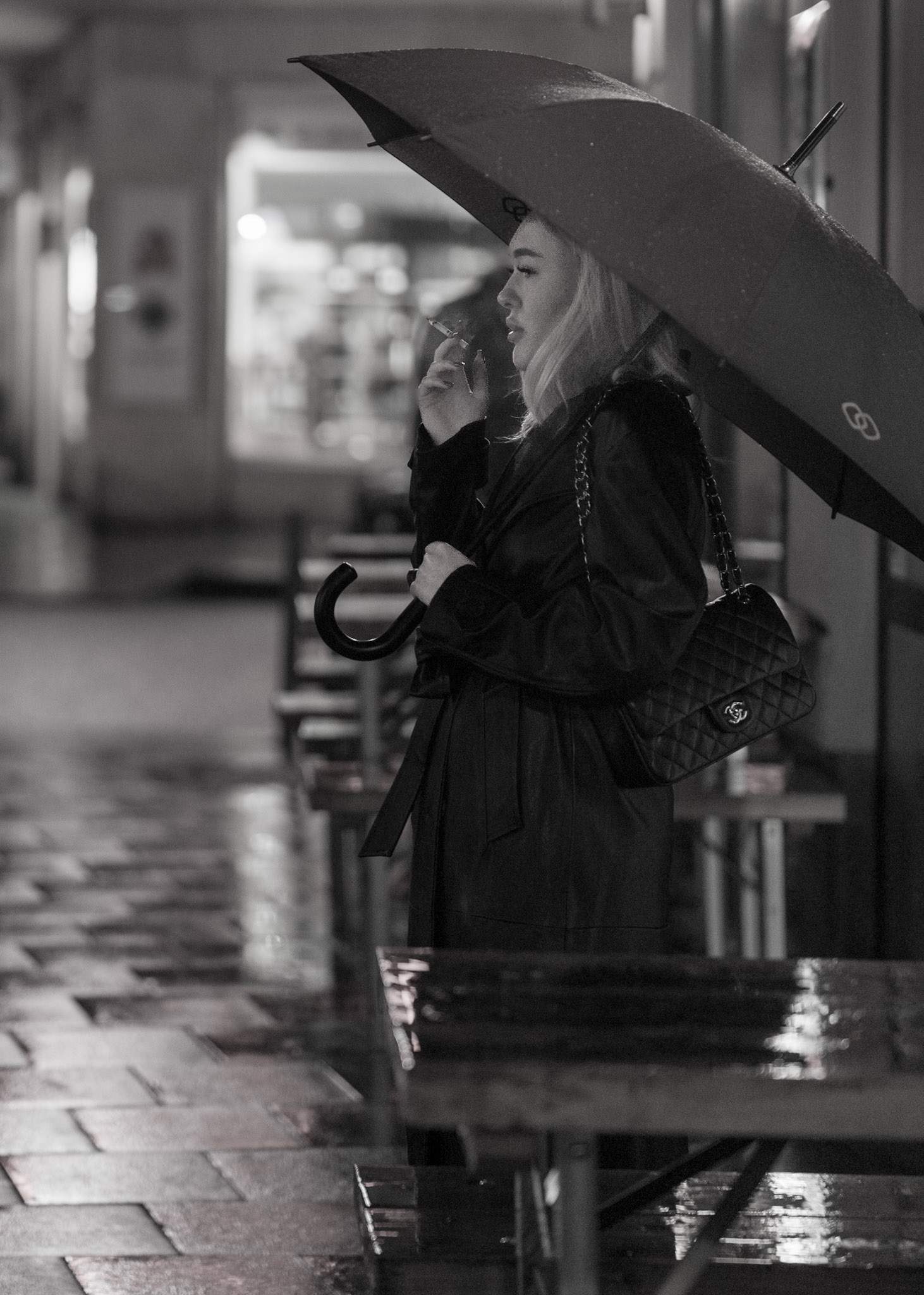 in the heart of munich's old town, a moment is cloaked in rain and reflections. a young woman stands, a solitary silhouette under an umbrella, the night air carrying the whisper of her thoughts with the smoke that drifts away. she's a portrait of the city's nocturnal poetry, each drop of rain an echo of stories untold. the restaurant's lights spill out onto the wet cobblestones, framing her in a hushed glow, a quiet interlude in the evening's rhythm.