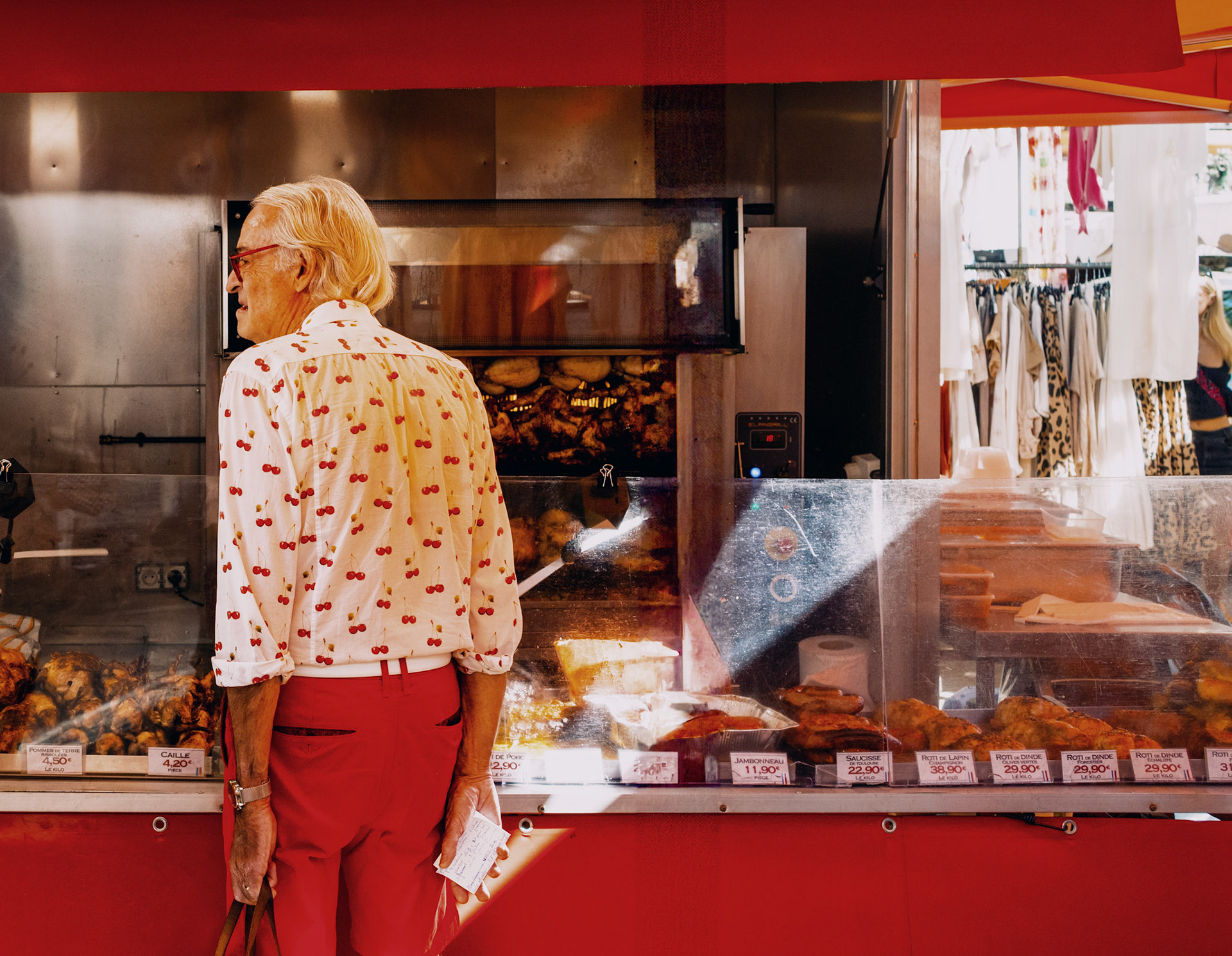 on a vibrant day at the place des lices market in saint tropez, the scene is an eclectic mix of sights and scents. an elderly man, adorned in red trousers, a shirt peppered with cherry prints, and red glasses, stands thoughtfully before a bustling rotisserie stand. the vivid red of his outfit harmonizes with the bright decor of the chicken grill, creating a striking visual contrast against the sizzling, golden chickens. this snapshot encapsulates a whimsical, yet poignant, interplay of life and culinary tradition, wrapped in the warm hues of a mediterranean afternoon.