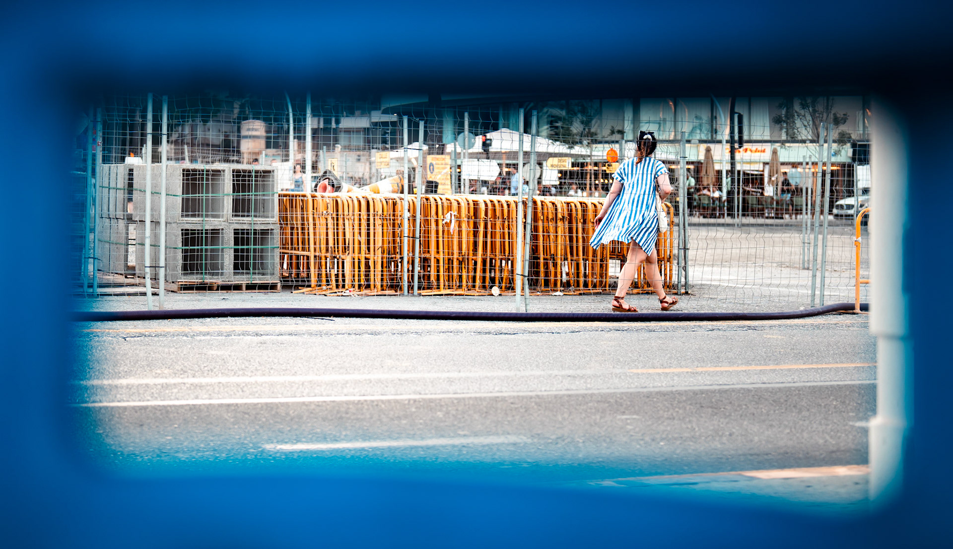 seen through a temporary fence, the scene contrasts the warm, flowing motion of a woman in her striped dress against the rigid, industrial layers of concrete and barriers on palma’s paseo marítimo. the construction has altered the landscape for two years now, yet life continues to weave through its interruptions. her bright presence highlights the resilience of movement in a place constantly evolving. even amidst the cold steel and unfinished paths, there’s warmth and softness.