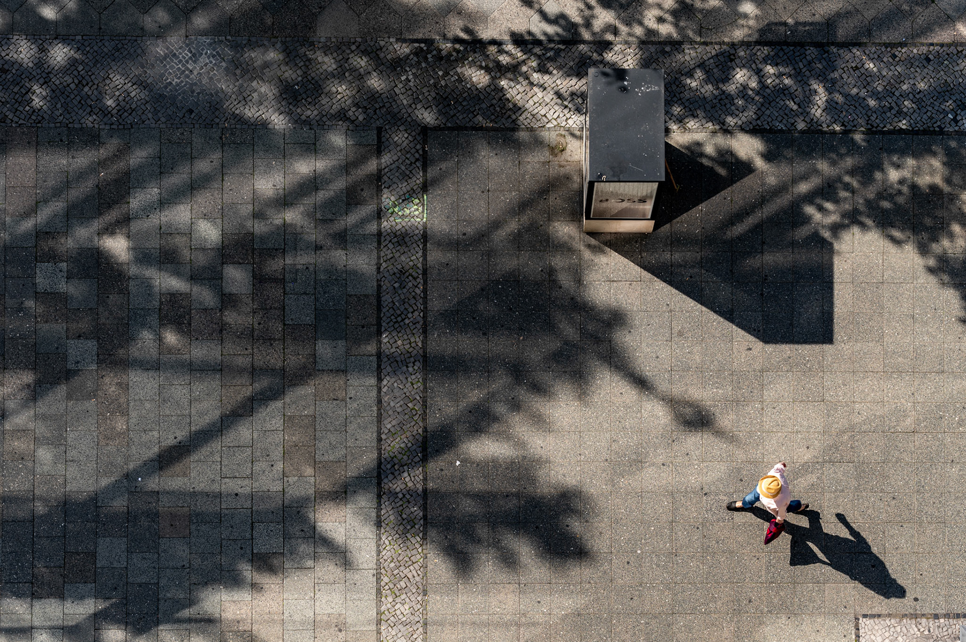 from above the bustling kurfürstendamm in berlin, this image captures a whimsical juxtaposition of past and present. a man in a straw hat walks purposefully, casting a long shadow that merges with the intricate patterns of the pavement and the abstract silhouettes of trees. this nod to classic street photography, where men in hats were once a common sight, plays with the elegance of tradition while firmly grounding itself in contemporary urban life. the interplay of light and shadow, structure and spontaneity, evokes a timeless narrative of the ever-evolving city.