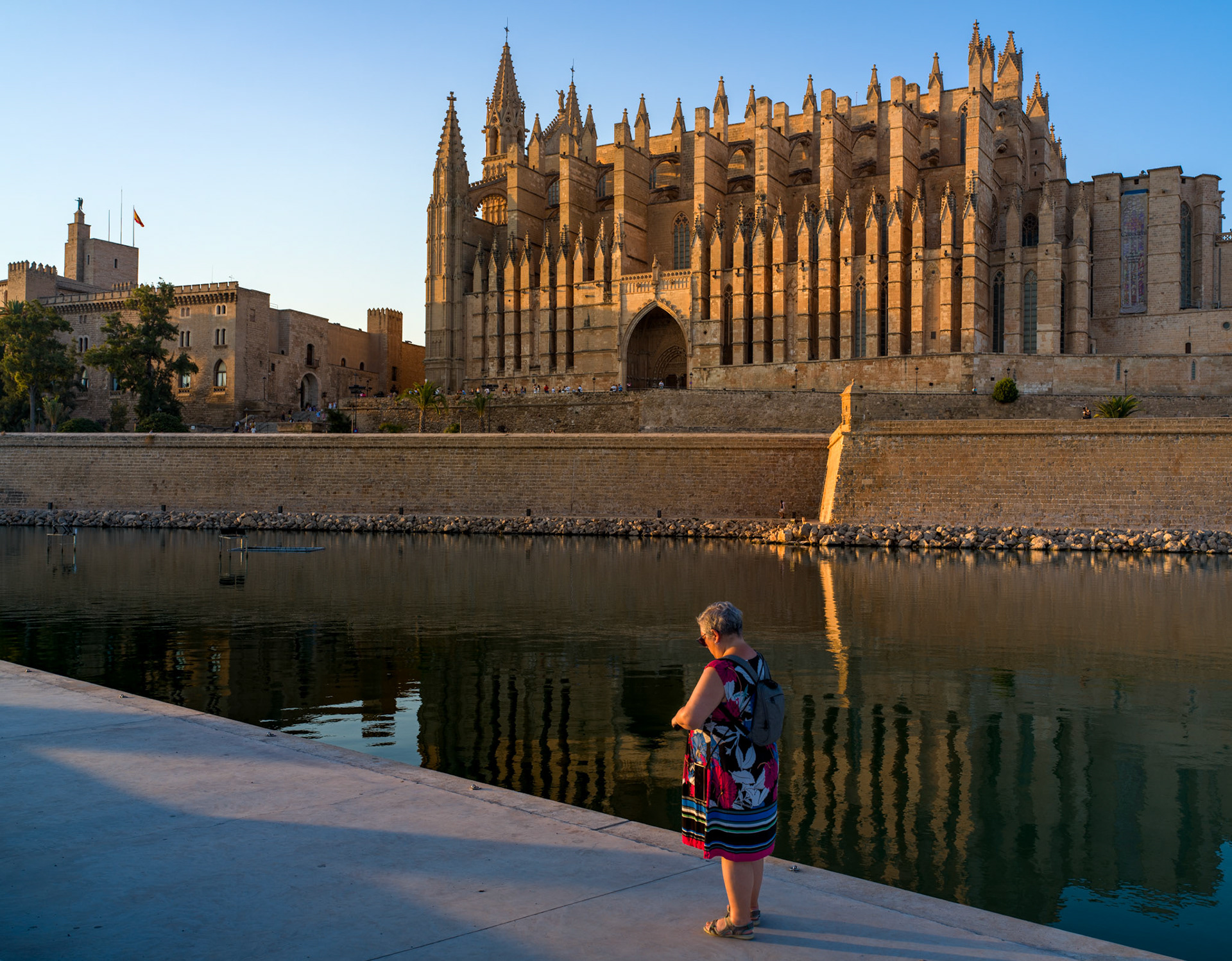 standing at the water's edge, she fiddles with a selfie stick, trying to capture the perfect memory. in the background, the grand silhouette of la seu cathedral dominates the scene, its sandstone walls glowing in the soft, golden light of the setting sun. the reflections ripple gently in the water, creating a serene mirror of both the iconic architecture and the fleeting moment of her quiet focus.