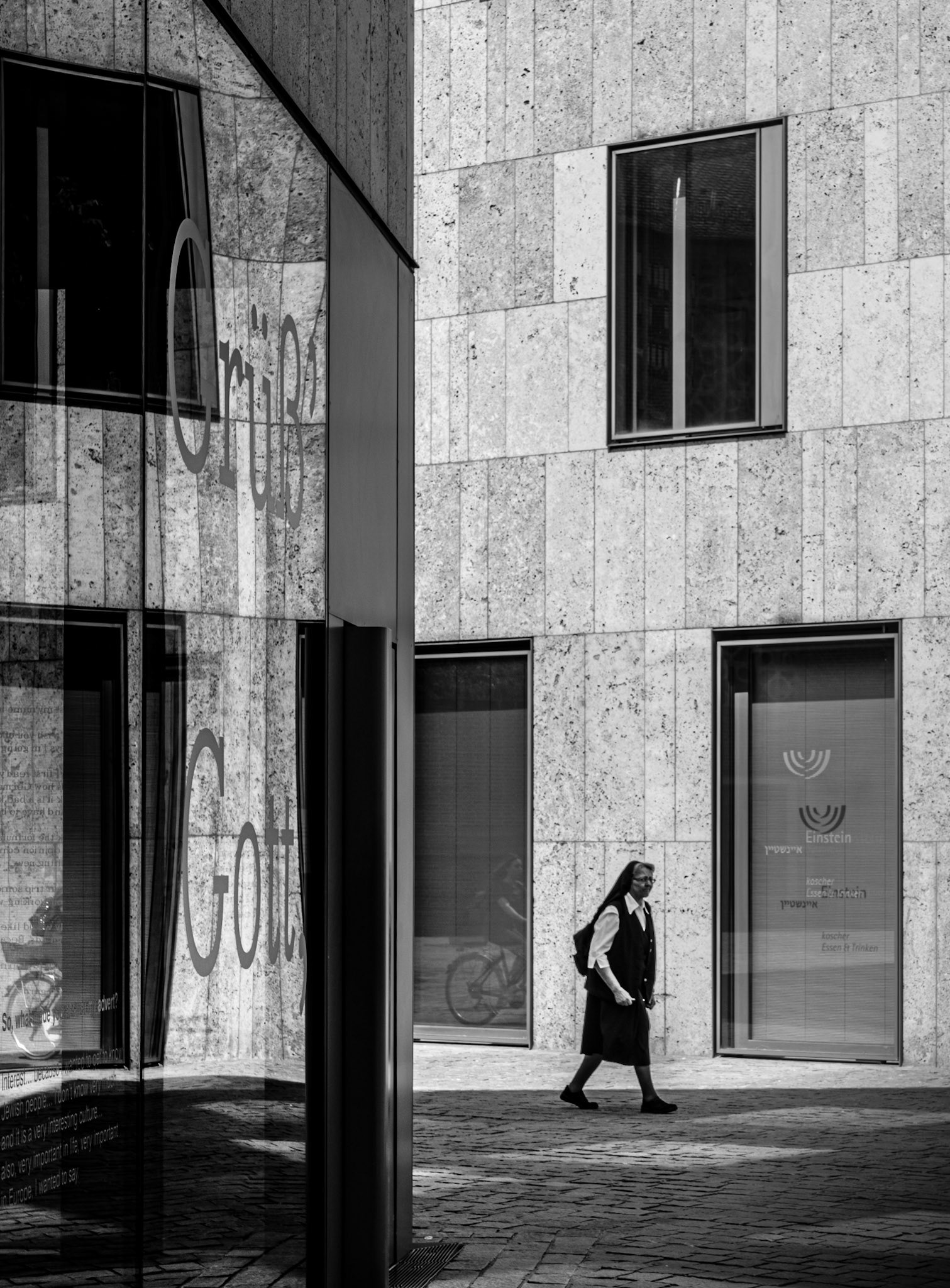in the quiet, reflective moments of an urban afternoon, a nun strolls past a modern facade, her presence a serene juxtaposition against the stark, minimalist architecture. the phrase "grüß gott" etched in the glass window mirrors her silent greeting to the world, blending tradition with contemporary life. the monochrome palette accentuates the timelessness of the scene, where the everyday intersects with the divine. it's a snapshot of quiet contemplation, capturing a fleeting moment of spiritual grace amidst the hustle and bustle of city life.