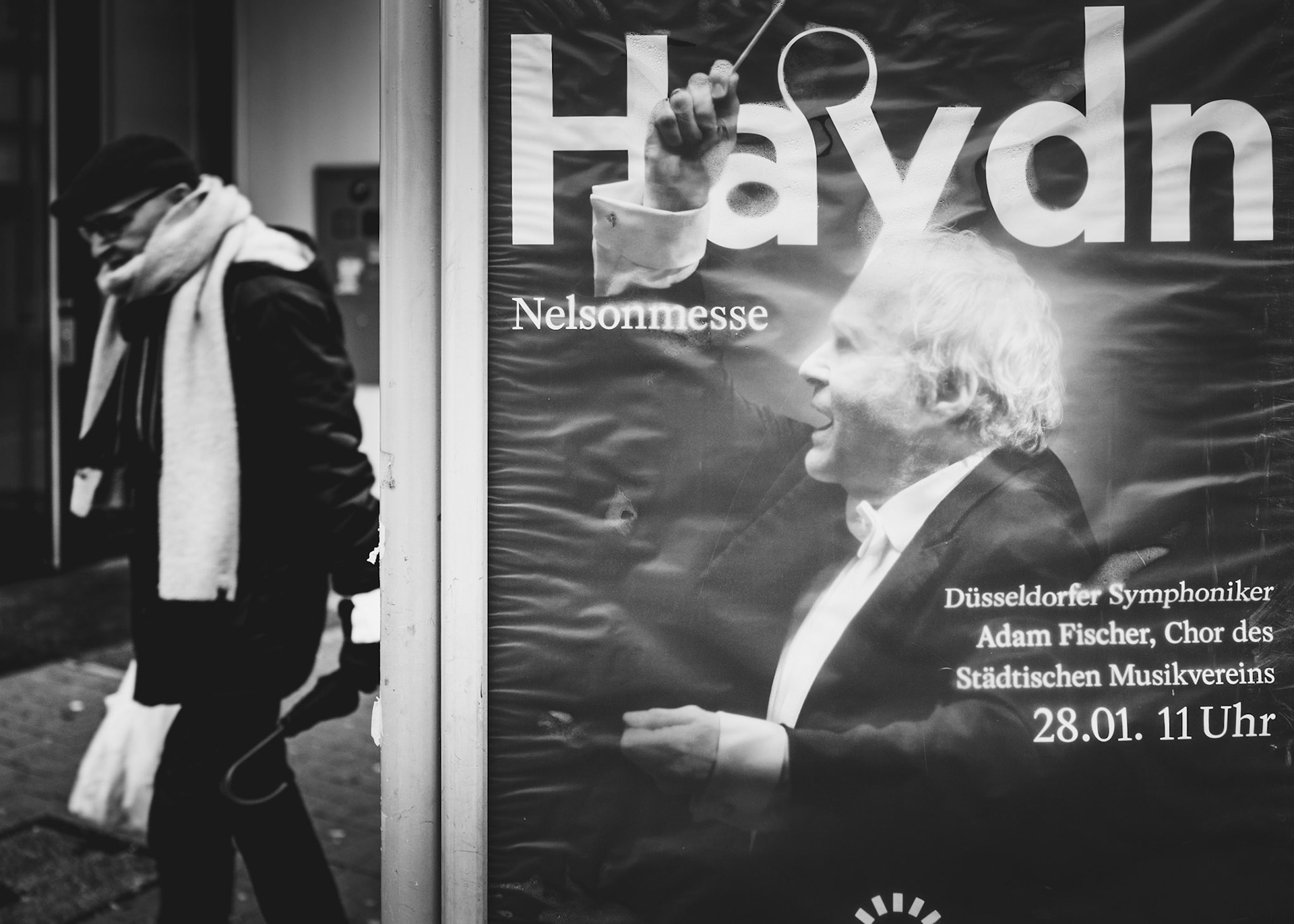 on a drizzly winter day in düsseldorf, a chance encounter between a passerby and a conductor's commanding image creates a striking scene. the man, wrapped in his winter attire, seems to be the focus of the conductor's intense energy, captured on the poster behind the glass. The rain adds a reflective sheen to the city's ambience, enhancing the serendipitous juxtaposition in this black-and-white photograph.