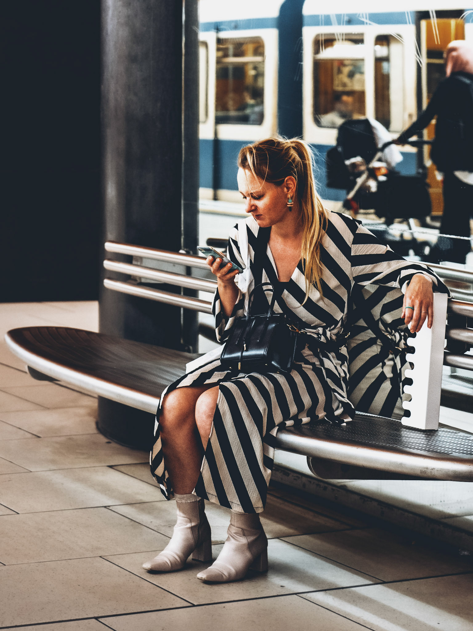amidst the city’s pulse, a moment of stillness. seated on a sleek metallic bench, the woman in bold black and white stripes becomes part of the geometry of the city. as she peers into her phone, the world moves behind her—a passing tram, fleeting shadows of strangers. the contrast between her stationary presence and the motion of the background creates a visual symphony, where patterns of stripes and urban lines merge into a narrative of quiet contemplation in the midst of the bustling city.