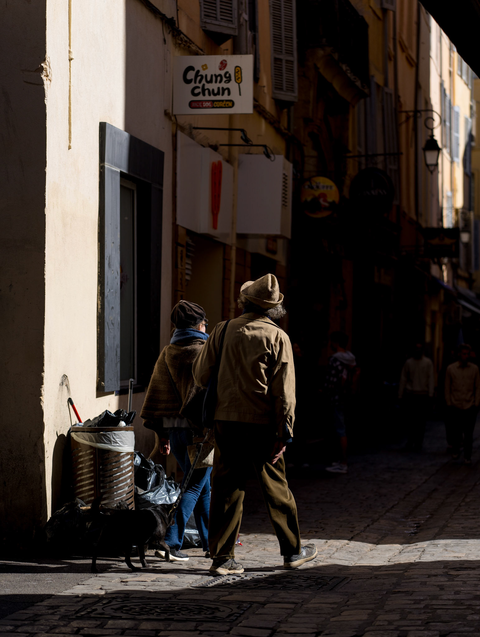 down the alleys of aix's old town, where the murmur of yesteryears clings to the facades, a couple with their dog weaves through a play of light and shadow. it's a living scene, where every detail whispers its own tale – the proud old walls, the modest heap of refuse, witness to the daily. the harsh contrast sketches a metaphor for life's intricate ballet of beauty and neglect, togetherness and solitude. they walk together, a small island of connection in the city's expansive story, framed by the chiaroscuro that conceals and yet reveals.