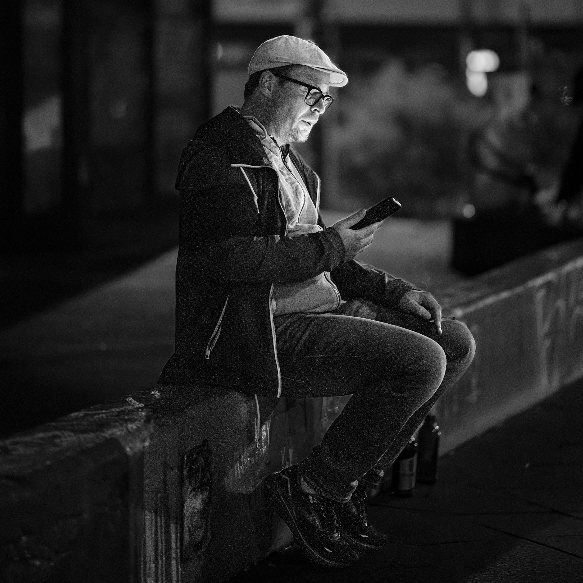 the soft glow of the phone reflects the quiet thoughts of the man sitting alone at night. the scene is still, the city hums in the background, yet here is a moment of calm, illuminated only by a small screen. the contrast between the darkened streets and his face, bathed in light, tells the story of modern solitude.