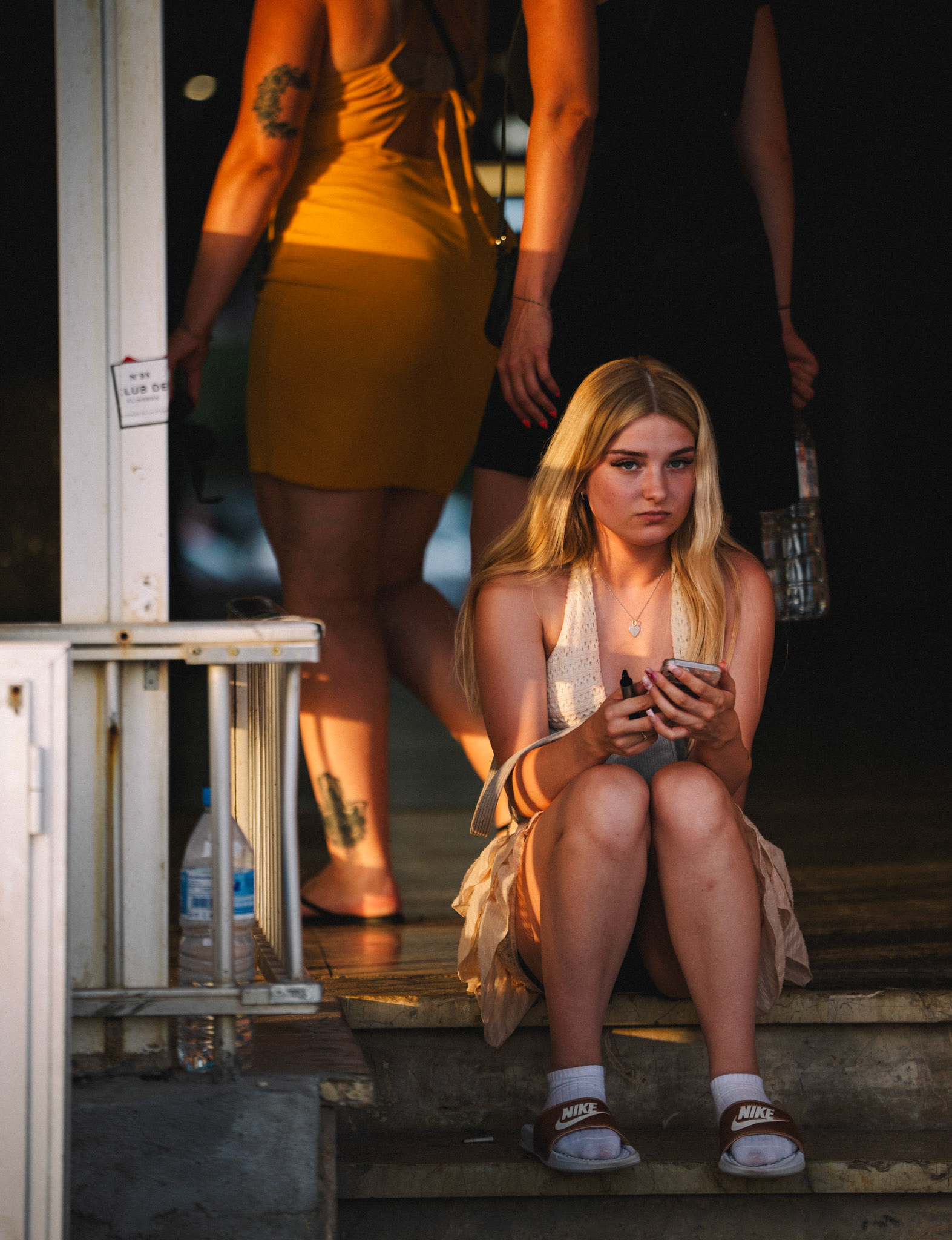 captured in the lively streets of el arenal, this image highlights a moment of quiet reflection. a young woman sits on the steps, her face touched with a hint of sadness as she looks down at her phone. around her, the world moves on—partygoers in vibrant dresses, full of energy and life, pass by, seemingly unaware of her solitude. the contrast between her stillness and the bustling background emphasizes the transient nature of emotions, where moments of loneliness can exist even in the midst of celebration.