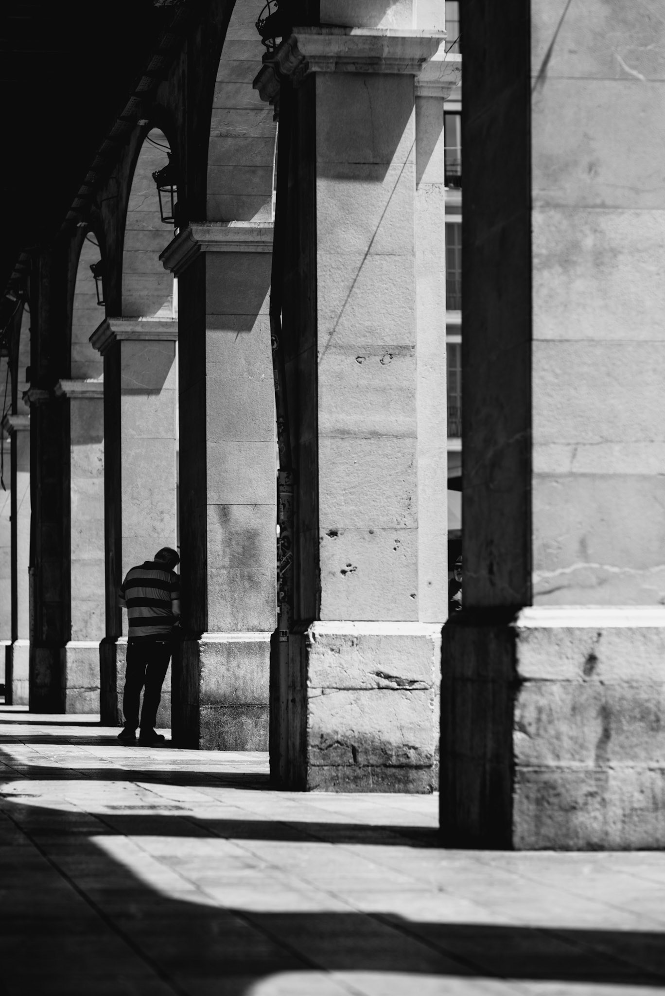 within the arches of plaça major, the interplay of light and shadow reveals a quiet scene. a solitary figure leans against the ancient stone pillar, lost in thought or perhaps a moment of solitude. the texture of the weathered stone tells tales of centuries past, while the shadows create a canvas of introspection. this black and white image captures the essence of silent conversations with history, a fleeting moment in the heart of palma de mallorca.