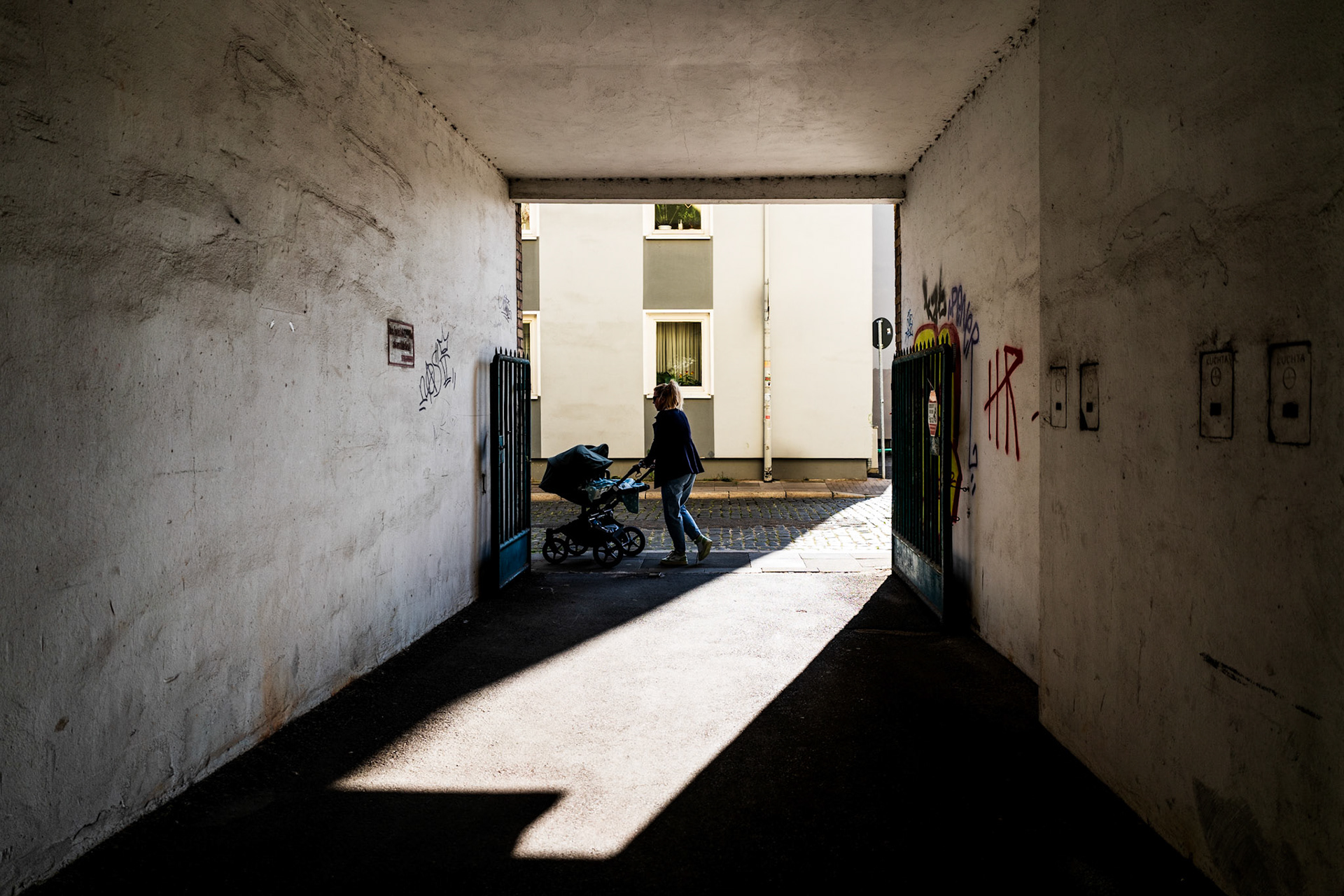 casting sharp shadows in a narrow passage, a figure pushes a stroller out into the brightness. graffiti adorns the weathered walls, adding a touch of urban art to the scene. this candid snapshot, taken in hannover, germany, contrasts the quiet routine of daily life with the city's textured backdrop.
