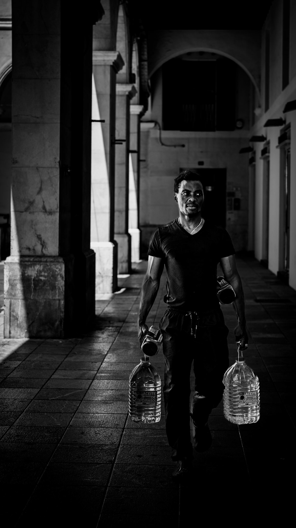 the man walks with determination, his hands gripping two heavy bottles, each one reflecting the weight of a daily grind. in his stride, there’s a quiet resilience against the sun's glare and the shadows of the plaza mayor in palma. the play of light and shadow, the stark contrast between his dark shirt and the pale stone archways, creates a striking portrait of strength and endurance. the scene tells a story of movement, purpose, and the simple, relentless rhythms of urban life. there’s an honesty here — an unfiltered moment caught in the passing of time, where every step feels deliberate, every shadow meaningful.