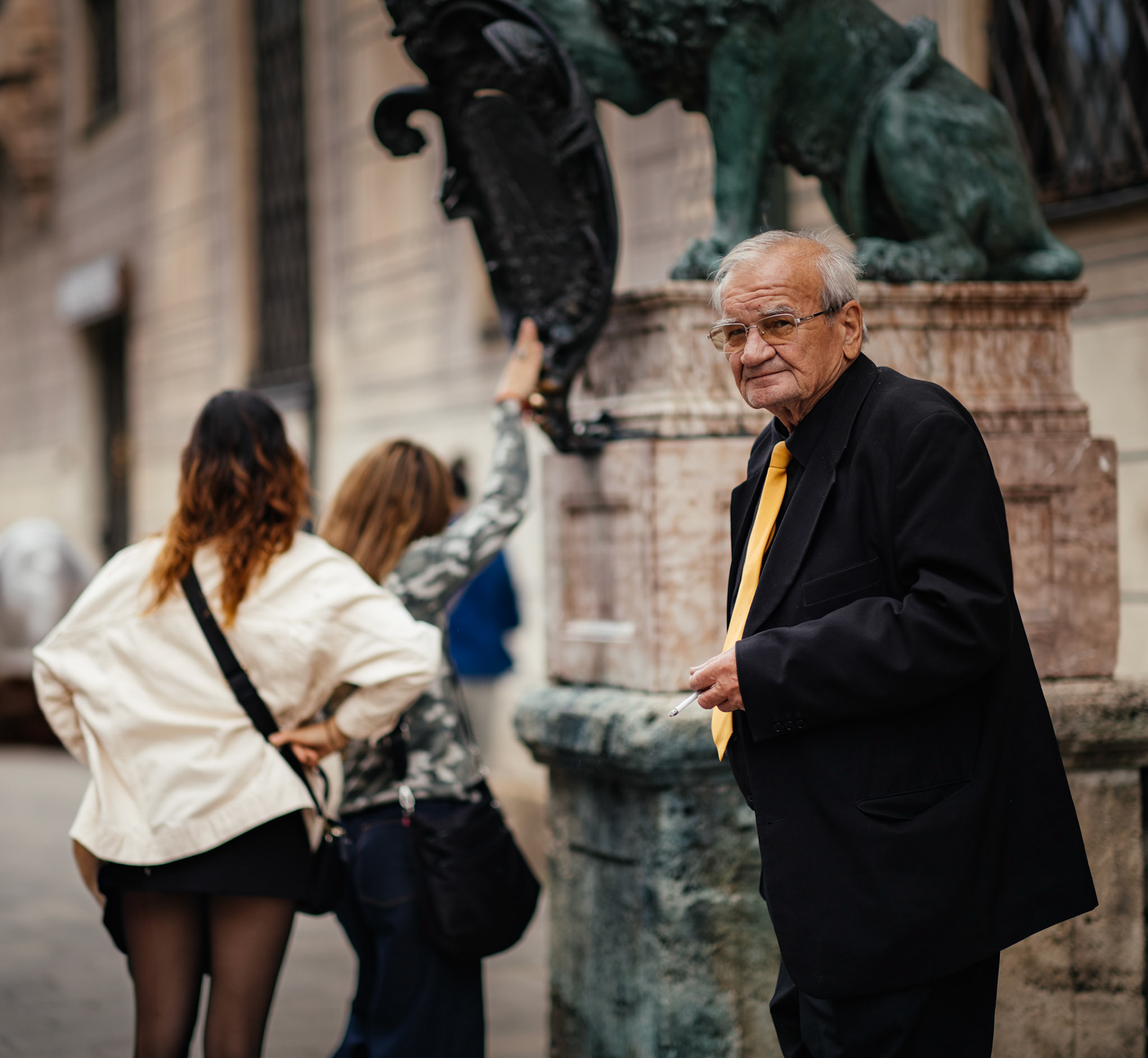 in front of the historic hofgarten in munich, an elderly man, dressed in a dark suit with a striking yellow tie, stands with an air of quiet vigilance. he appears to be the guardian of this place, observing the tourists who reach out to touch the lion statue, a gesture believed to bring good fortune. the contrast between the bustling curiosity of the visitors and the calm, watchful demeanor of the man creates a scene rich with storytelling. the timelessness of the location is echoed in the guardian's presence, symbolizing the silent keepers of tradition amidst the ever-changing flow of life.